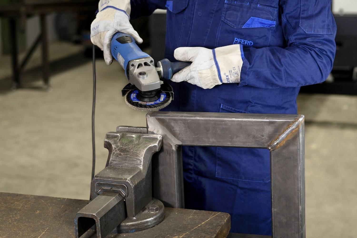 Eine Person in Arbeitskleidung hält einen Winkelschleifer und bearbeitet ein Metallstück in einer Werkstattumgebung.