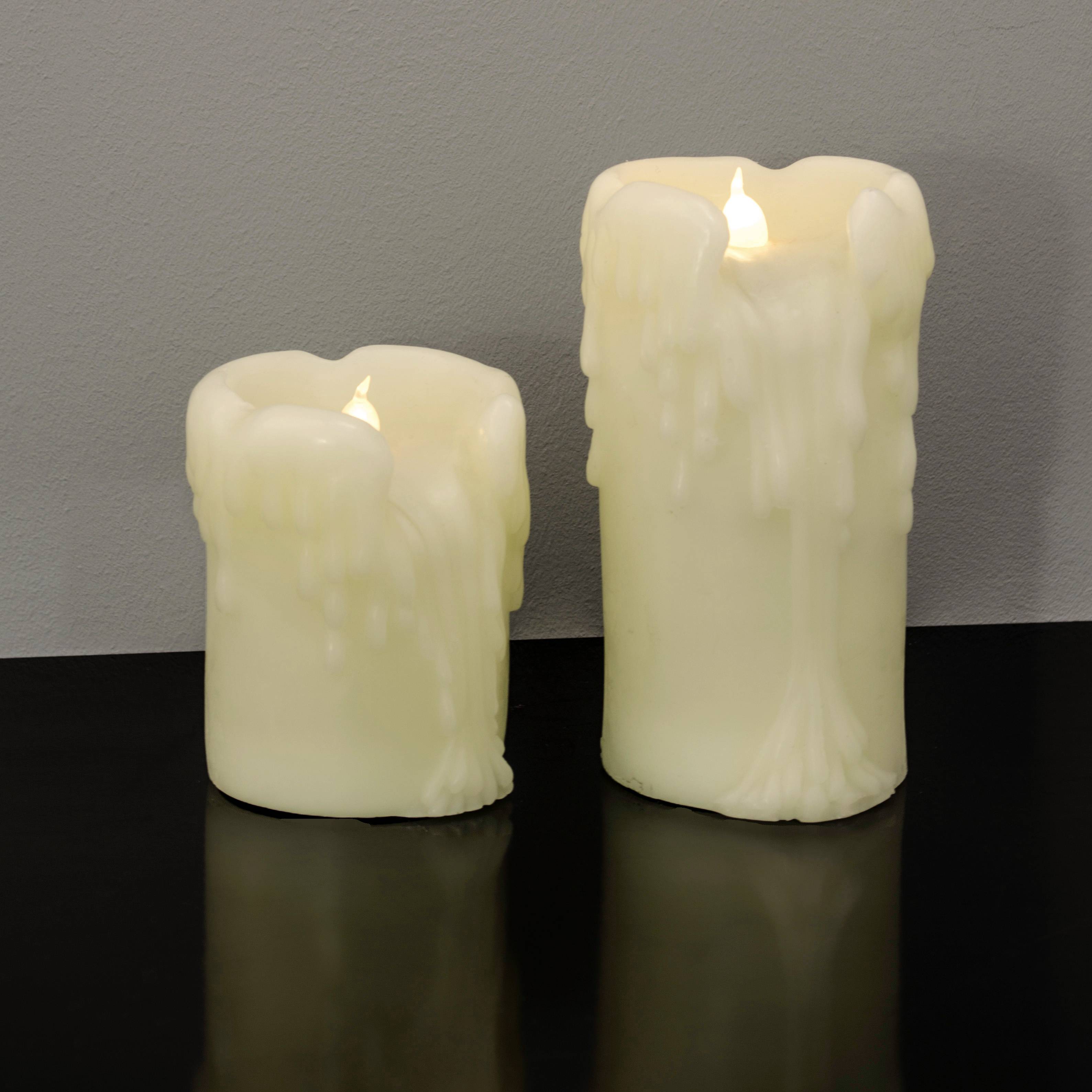 Zwei unterschiedlich große, weiße Kerzen mit flackernden Flammen, auf einem Tisch vor einer grauen Wand stehend. Die Kerzen wirken echt, sind aber unecht.