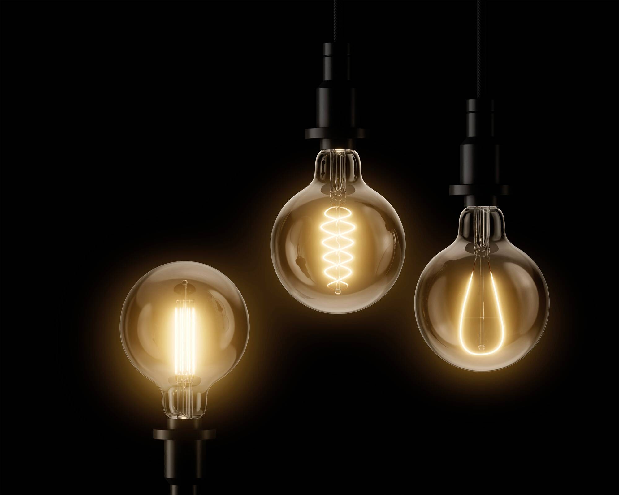 Drei Glühbirnen hängen vor einem schwarzen Hintergrund, jede mit unterschiedlichem Glühfaden-Design, und leuchten warm.