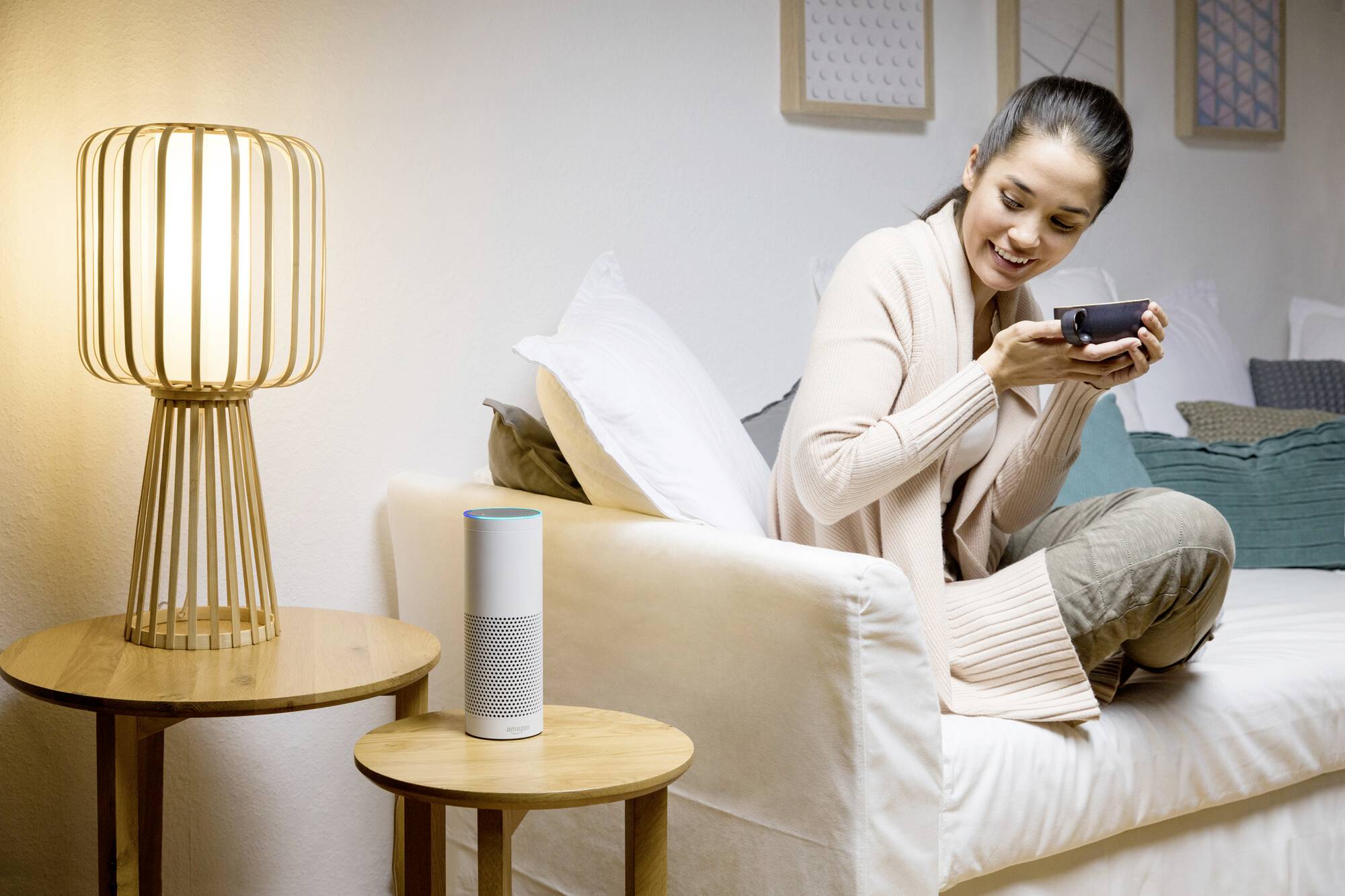 Eine Frau sitzt auf einem Sofa und trinkt aus einer Tasse. Neben ihr steht ein intelligenter Lautsprecher auf dem Tisch. Sie wirkt entspannt.