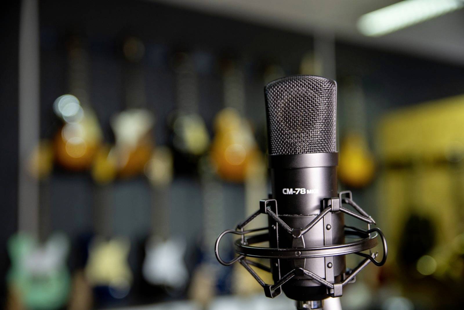 Ein Mikrofon im Vordergrund, unscharfe Gitarren im Hintergrund. Szenerie deutet auf ein Musikstudio oder Proberaum hin.