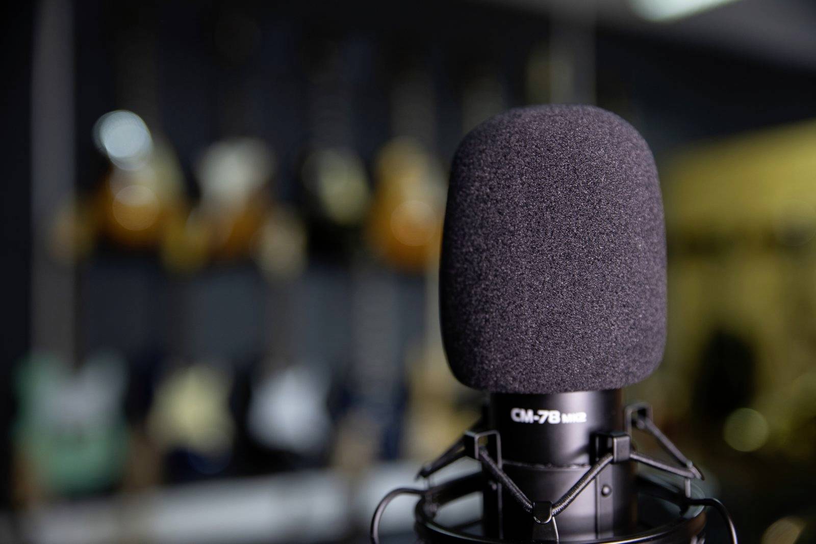 Ein schwarzes Mikrofon fokussiert im Vordergrund, unscharfer Hintergrund mit verschwommenen Gitarren an der Wand. Studioumgebung.