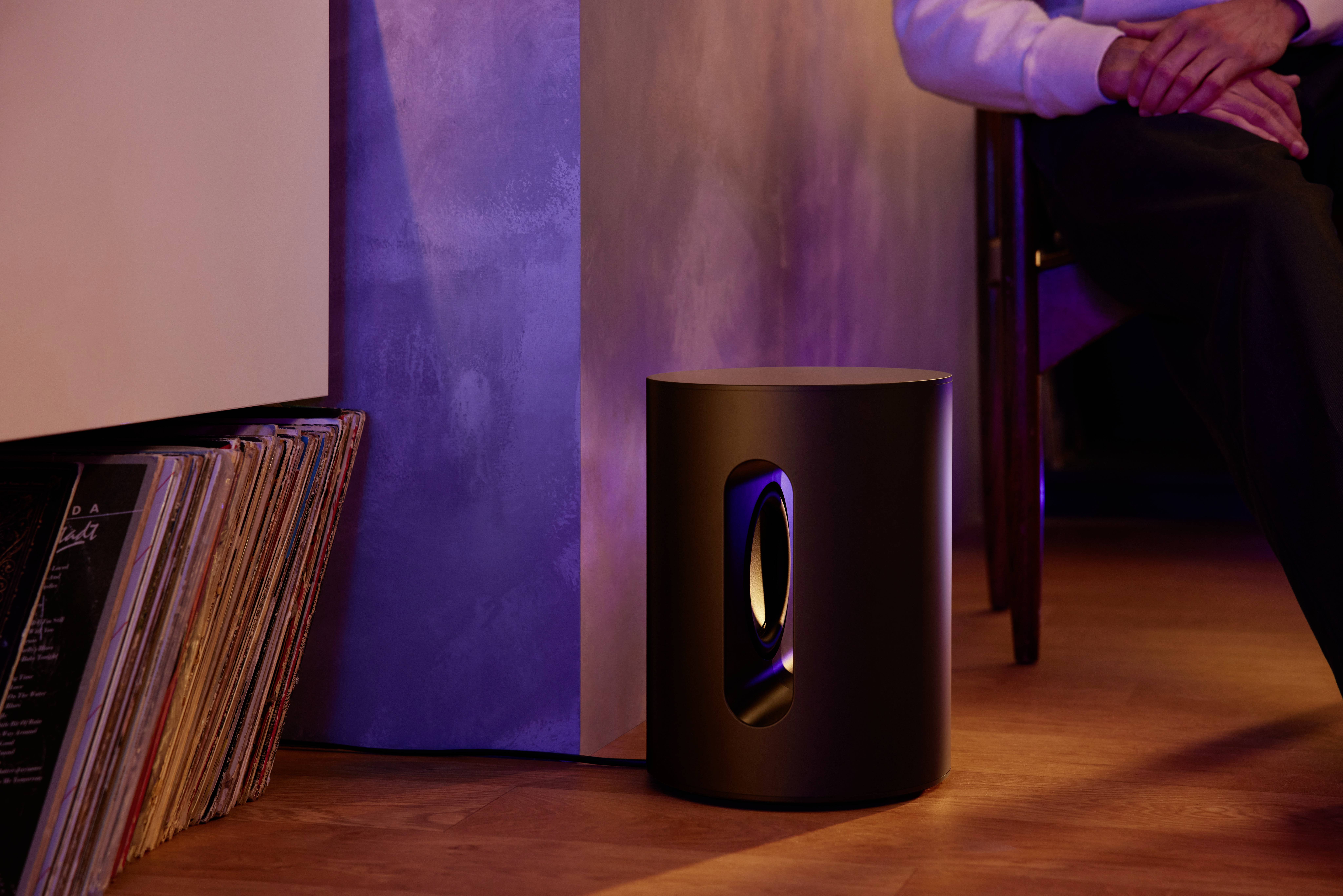 Ein schwarzer Lautsprecher steht auf einem Holzfußboden neben einer Wand mit Vinylplatten. Eine Person sitzt daneben.