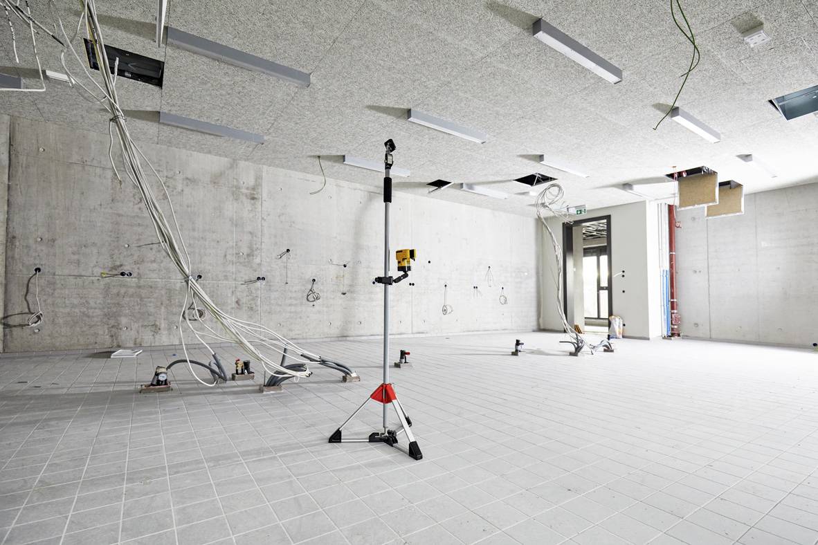 Baustelle in einem Innenraum mit freiliegenden Kabeln, einem Baustativ und unvollendeten Deckenarbeiten. Keine Arbeiter sichtbar.