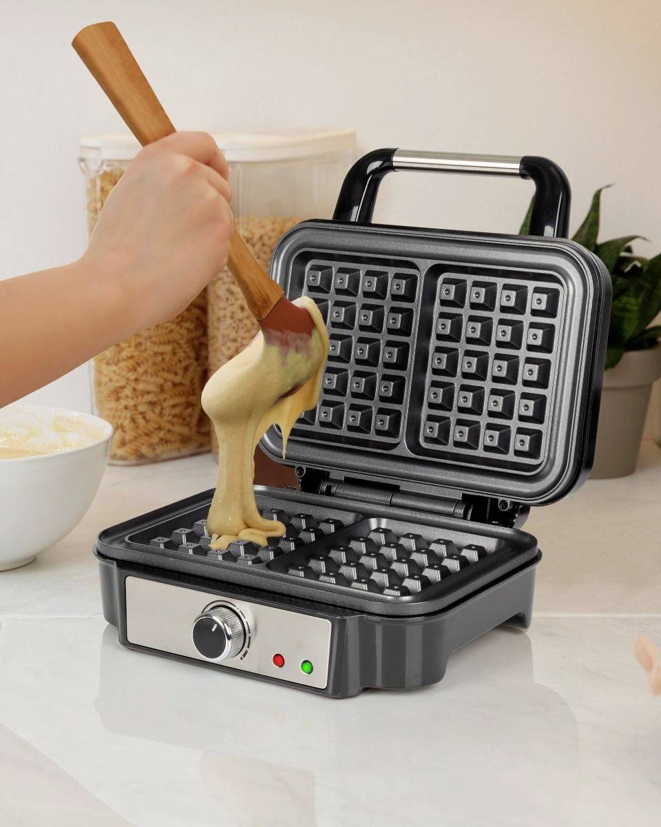 Eine Hand gießt Teig in ein schwarzes Waffeleisen auf einer weißen Küchenarbeitsplatte. Im Hintergrund sind Zutaten zu sehen.