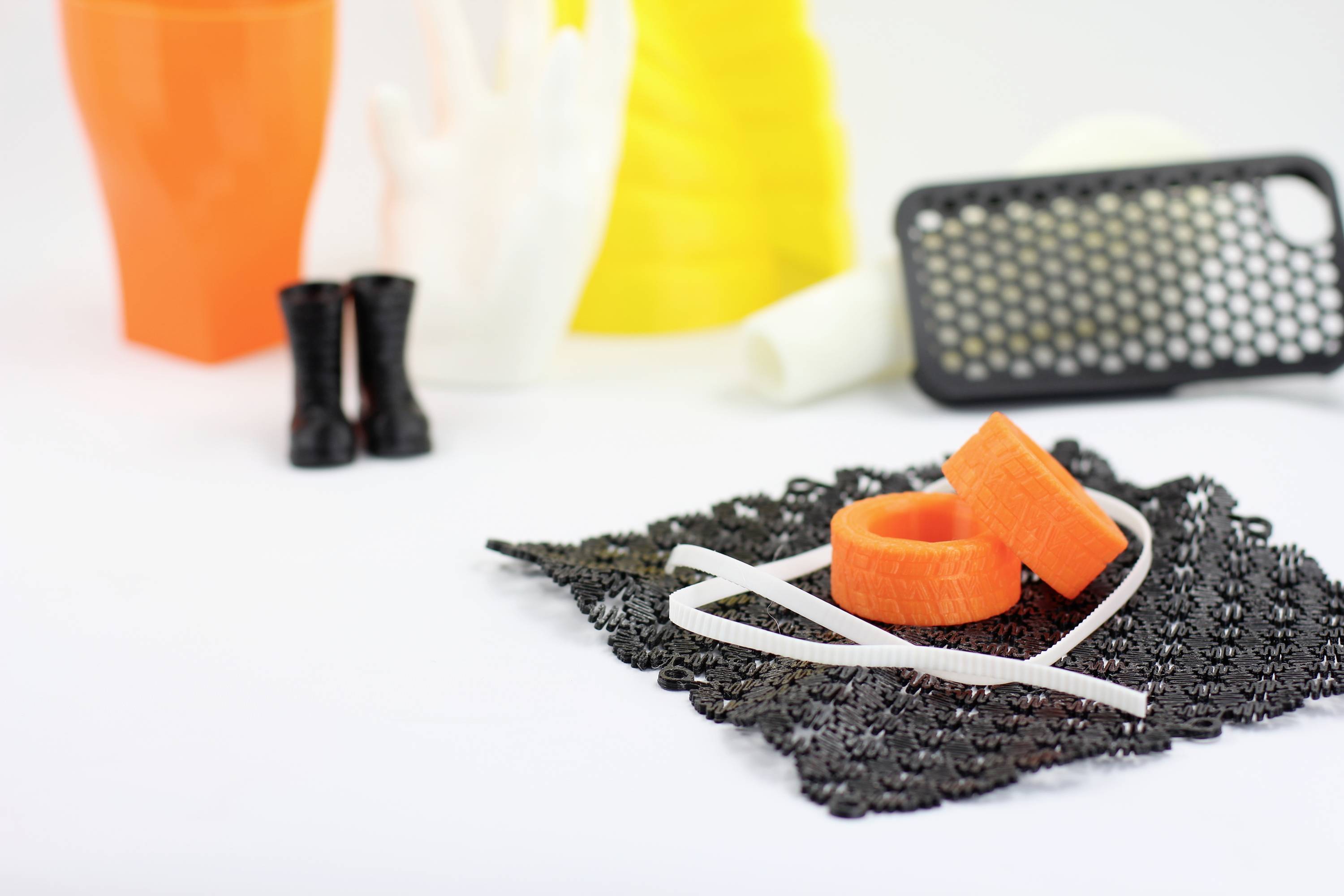 Mehrere 3D-gedruckte Objekte in verschiedenen Farben und Formen auf einer weißen Oberfläche; im Vordergrund ein schwarzes Netz mit orangefarbenen Ringen.