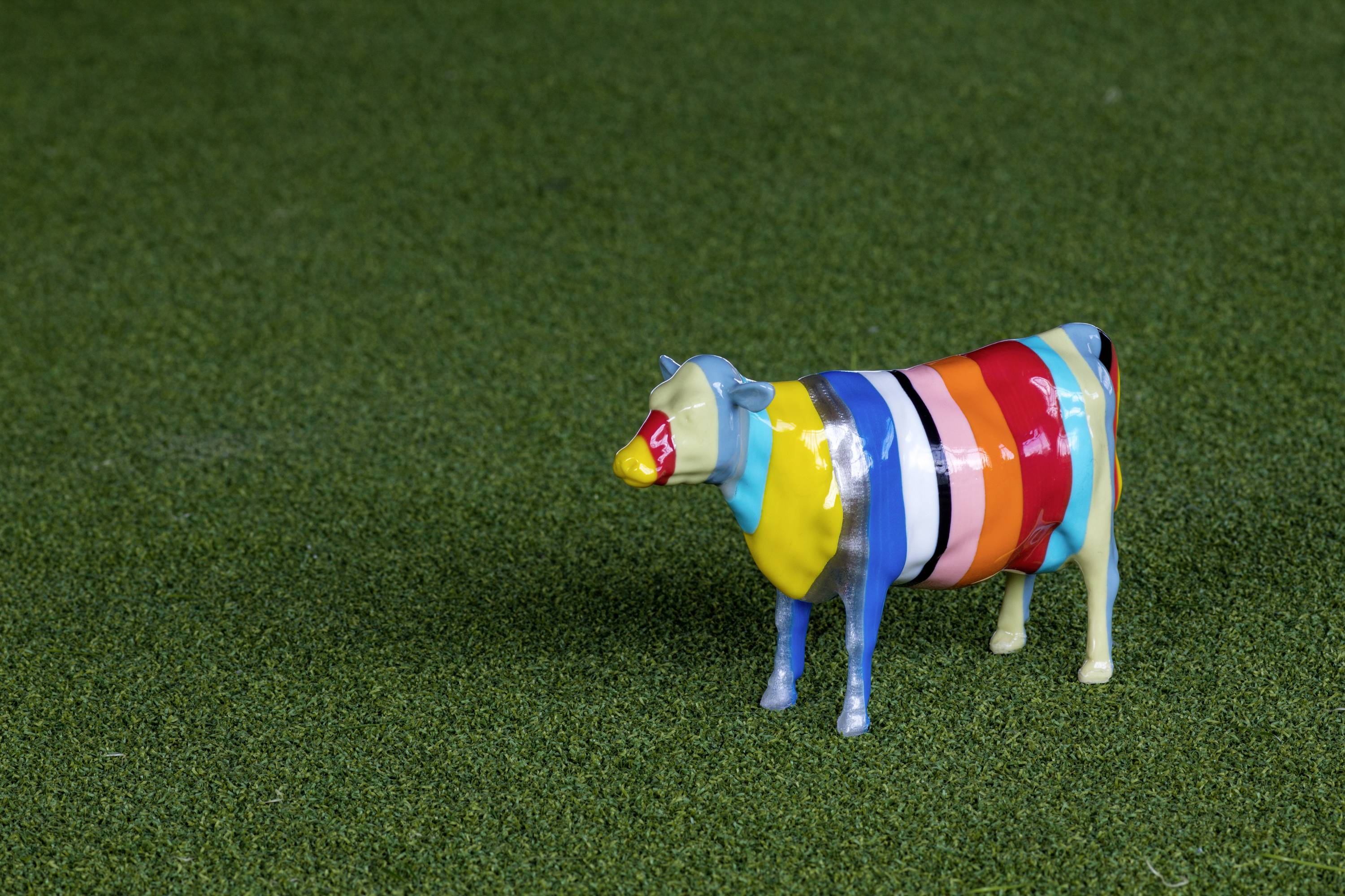 Eine kleine, bunt gestreifte Plastikfigur einer Kuh steht auf grünem, grasähnlichem Untergrund.