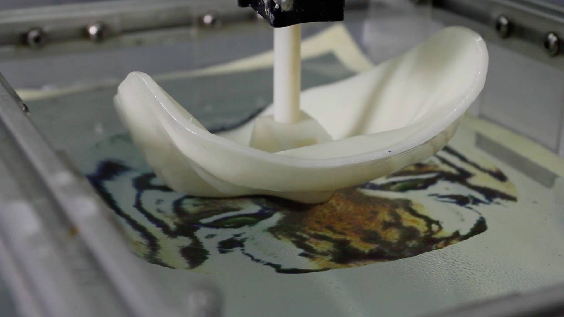 Ein 3D-Drucker formt ein geschwungenes Objekt. Im Hintergrund ist ein Bild eines Tigers auf einem Bildschirm zu sehen.