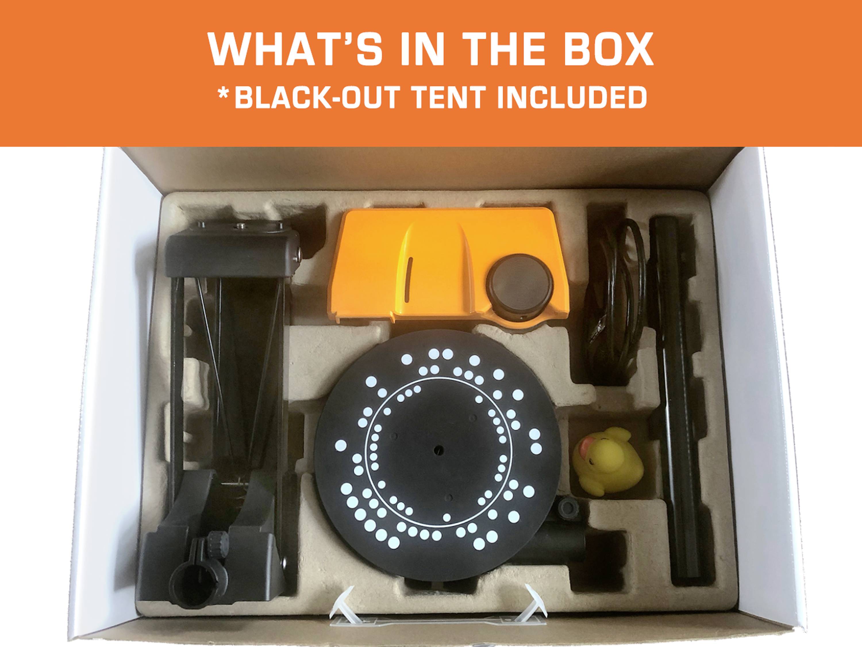 Kartonschachtel mit Inhalt: schwarze Leuchte, orangefarbenes Gerät, Kabel, Stativ und gelbe Ente. Text: 'WHAT'S IN THE BOX *BLACK-OUT TENT INCLUDED'.