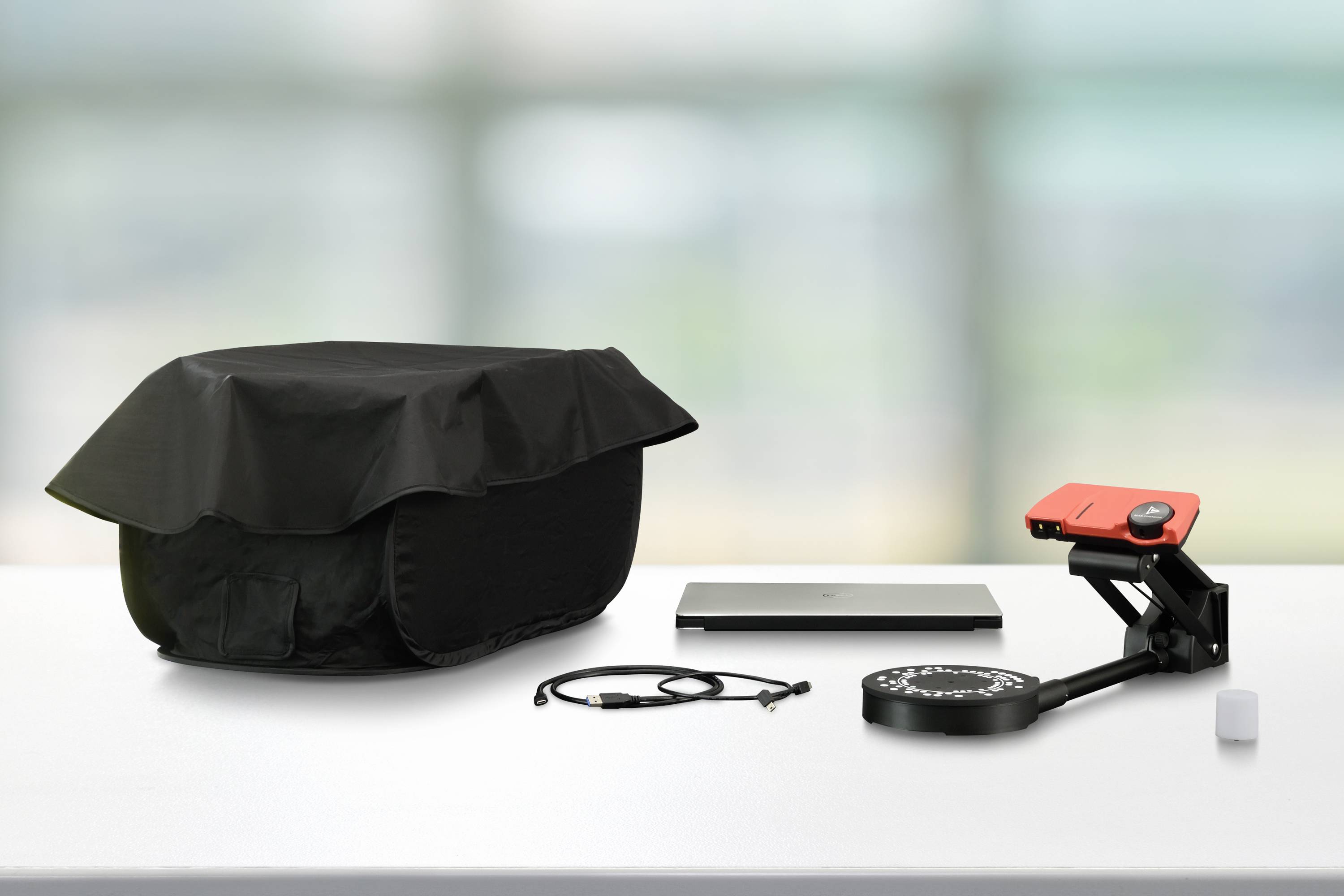 Ein Laptop, ein Projektor mit roter Abdeckung und ein schwarzer Schutzüberzug auf einem Tisch in einem hellen Raum.