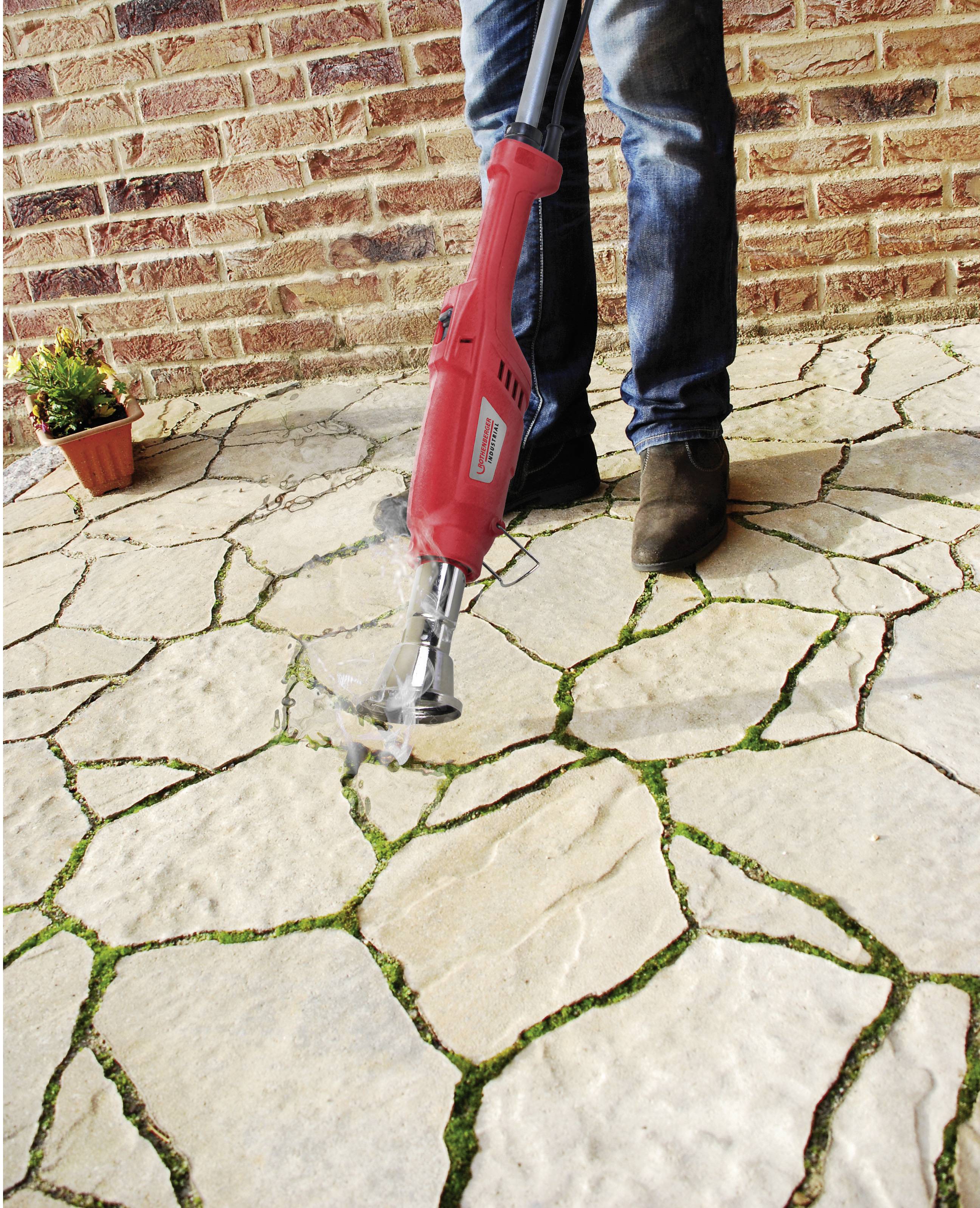 Eine Person entfernt mit einem roten Gerät Unkraut aus den Fugen eines gepflasterten Steinwegs vor einer Backsteinmauer.