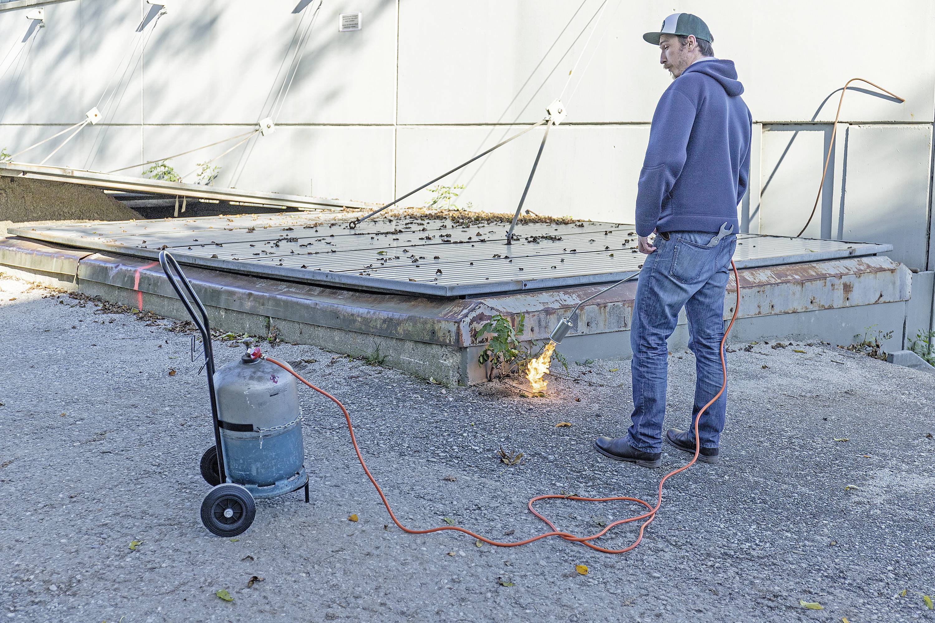 Ein Mann in Freizeitkleidung benutzt einen Gasbrenner, um etwas neben einem Gebäude zu erhitzen. Eine Gasflasche steht auf einem Wagen.