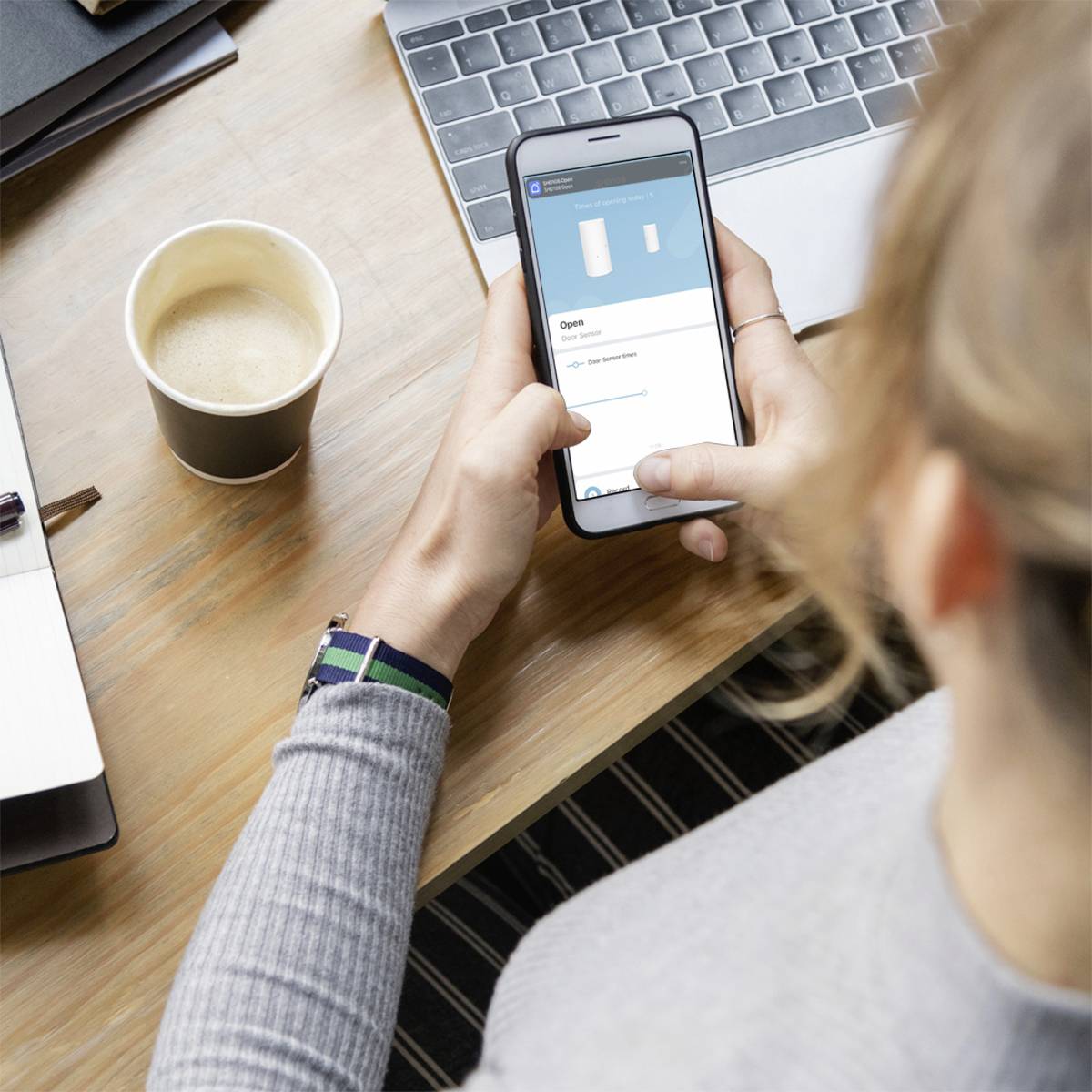 Eine Person hält ein Smartphone und betrachtet eine App, daneben ein Laptop und ein Becher Kaffee auf einem Holztisch.