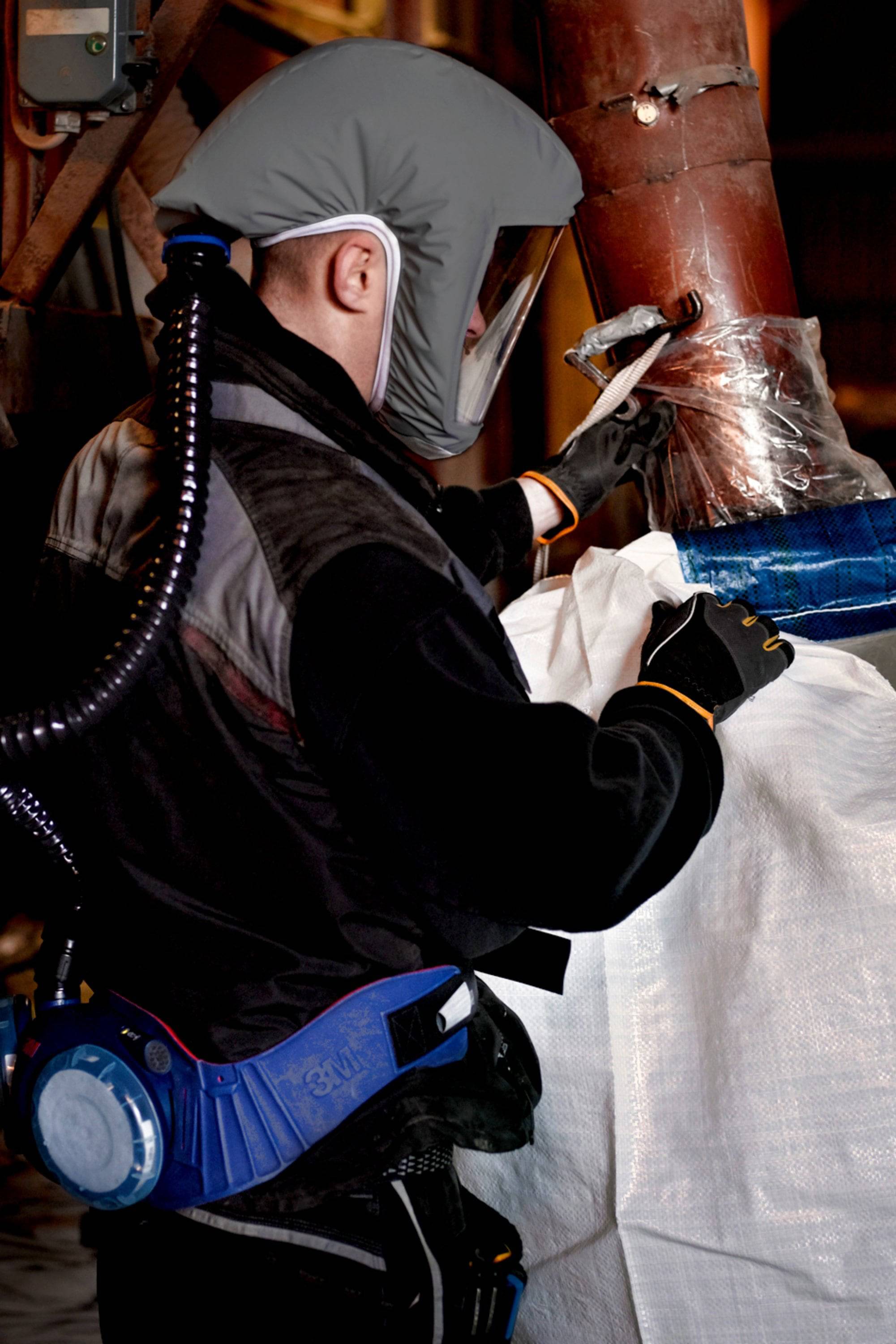 Eine Person mit Schutzkleidung und Atemgerät arbeitet an einem Rohr, das mit Plastikfolie umwickelt ist, in einer industriellen Umgebung.
