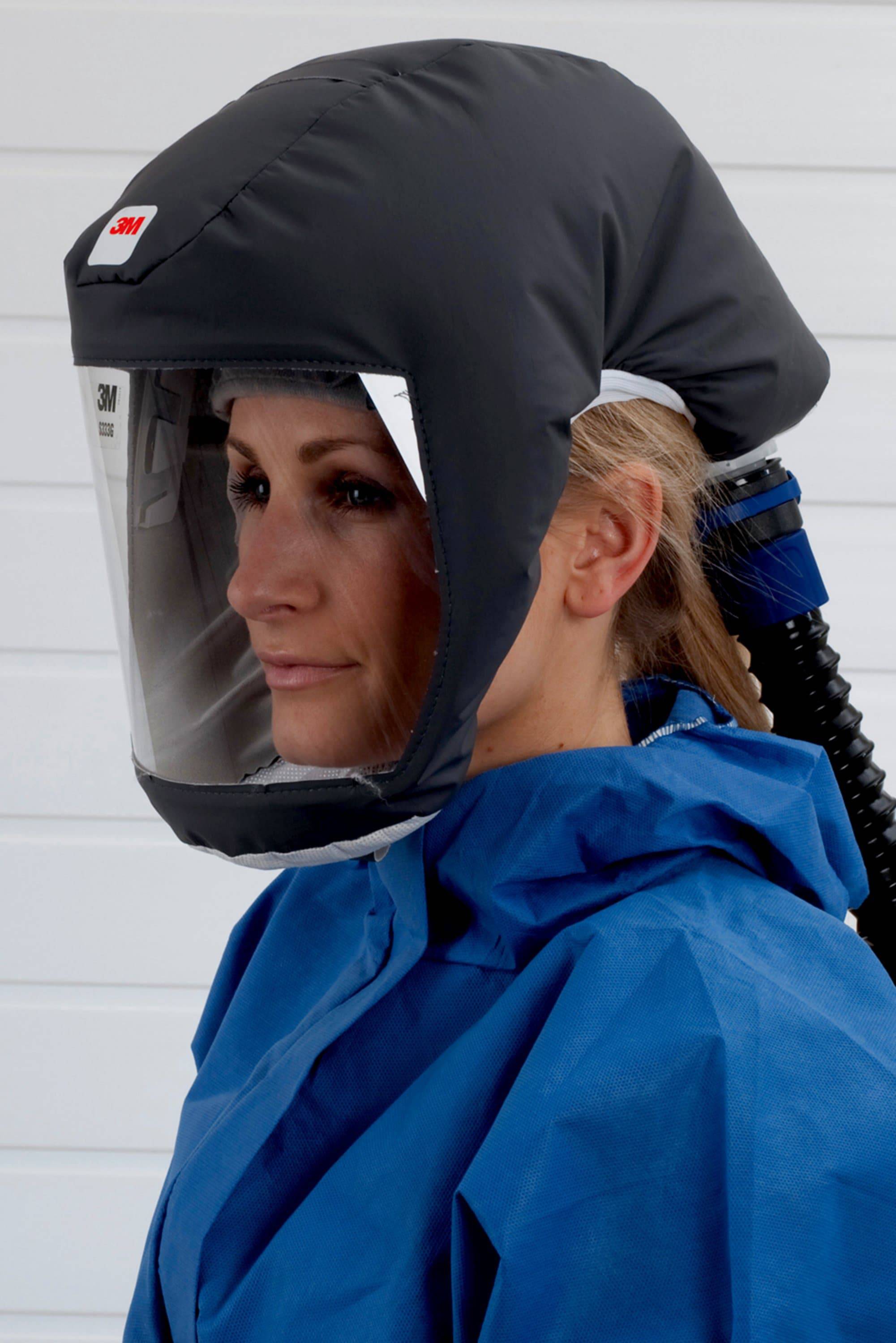 Eine Person trägt eine Schutzhaube mit Visier und Atemschutz, gekleidet in einem blauen Schutzanzug, stehend vor einem weißen Hintergrund.