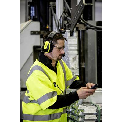 Mann in gelber Warnweste und Schutzbrille, arbeitet an einem Gerät in einer Fabrikumgebung, trägt Kopfhörer mit Mikrofon.