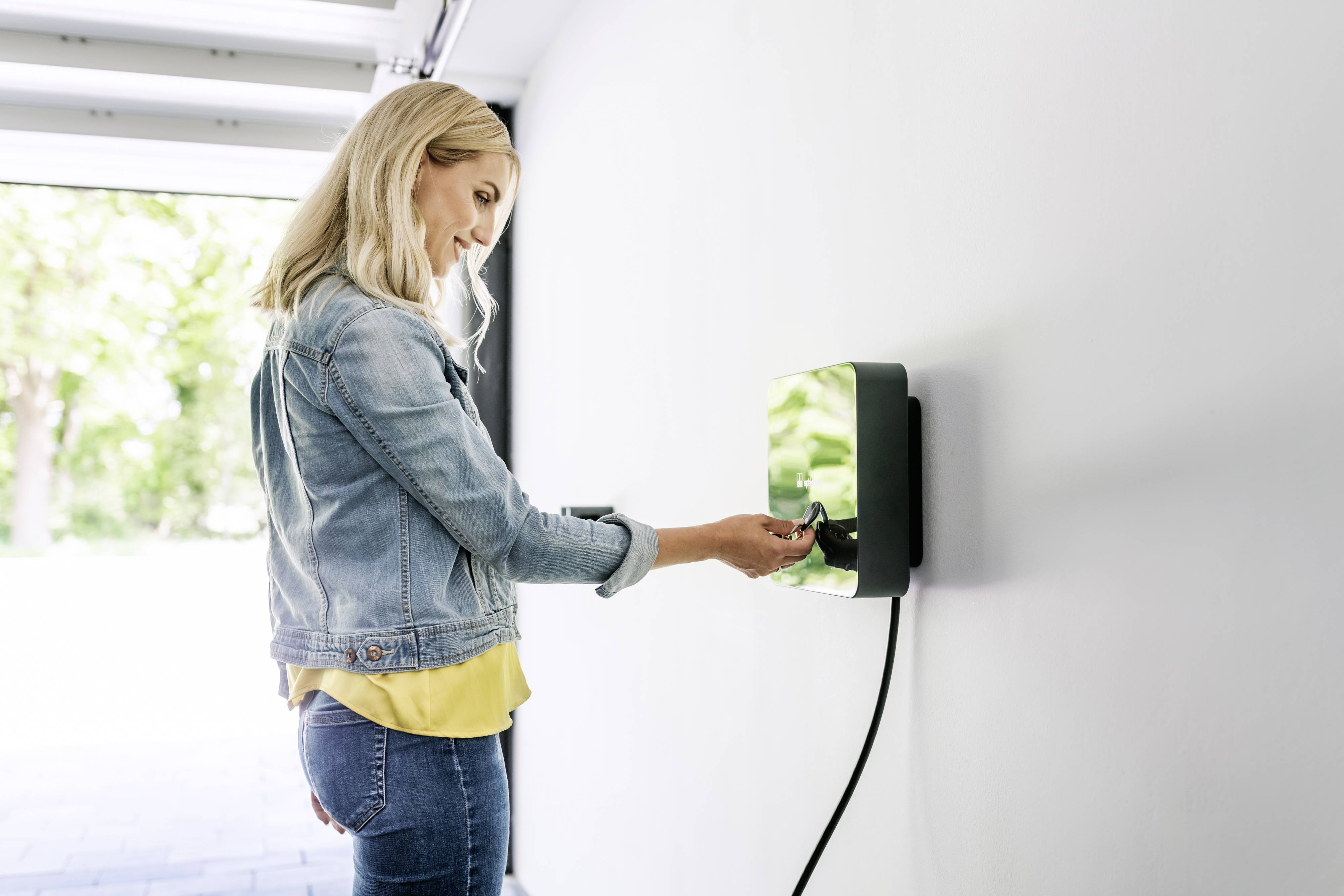 Eine Frau lädt ein Elektrofahrzeug an einer Ladestation in einer Garage auf. Sie trägt eine Jeansjacke und lächelt.