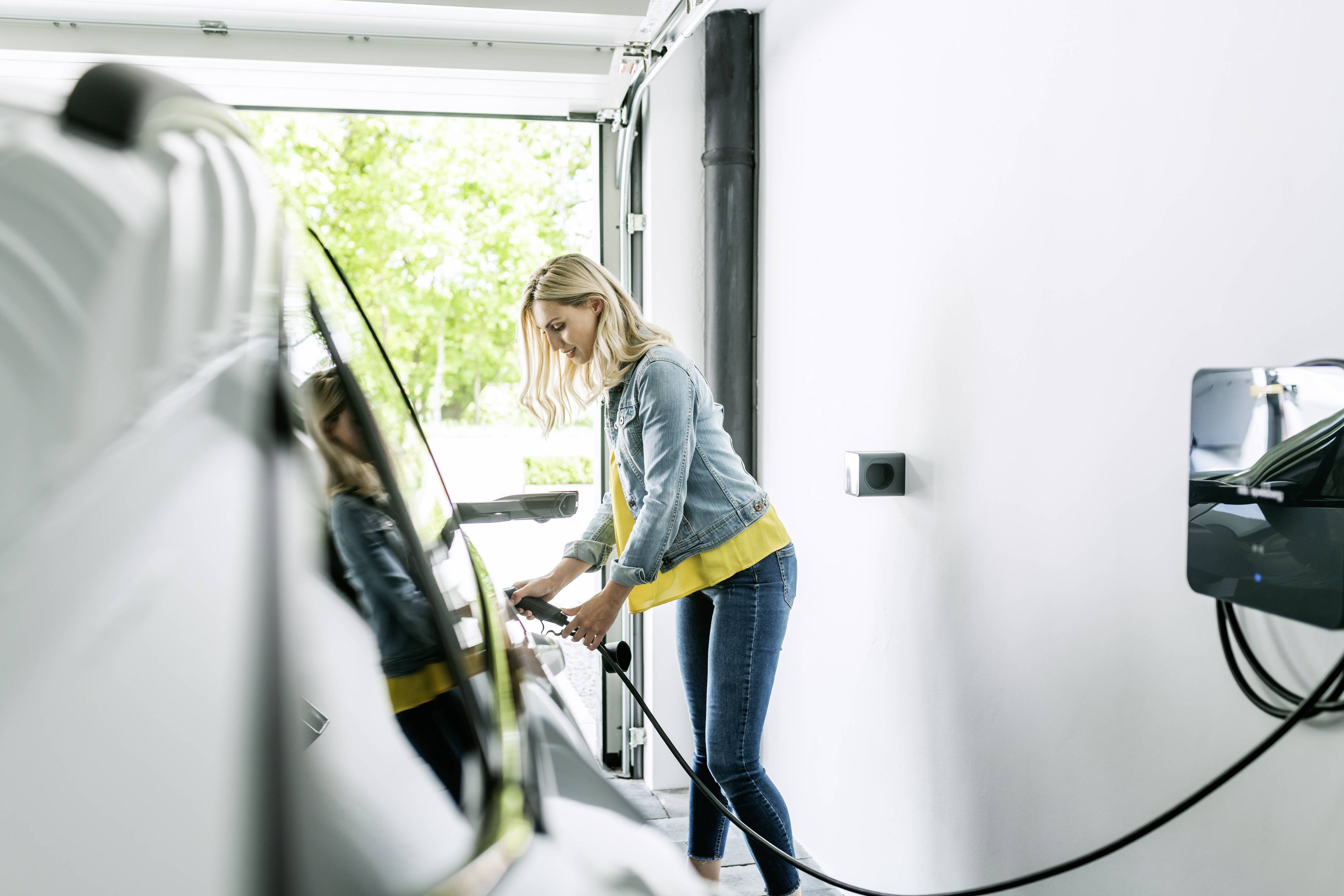 Eine Frau lädt ein Elektroauto in einer Garage auf. Sie hält das Ladekabel in der Hand, der Wagen ist nah an der Tür geparkt.