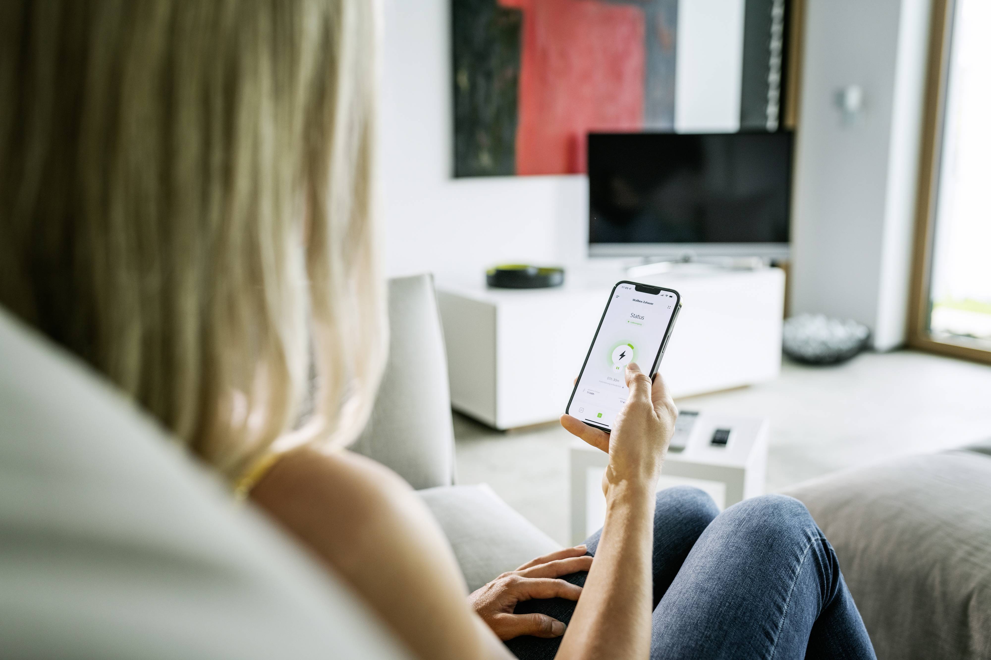 Eine Person sitzt auf dem Sofa und bedient ein Smartphone. Auf dem Bildschirm ist eine App mit einem grünen Häkchen zu sehen.