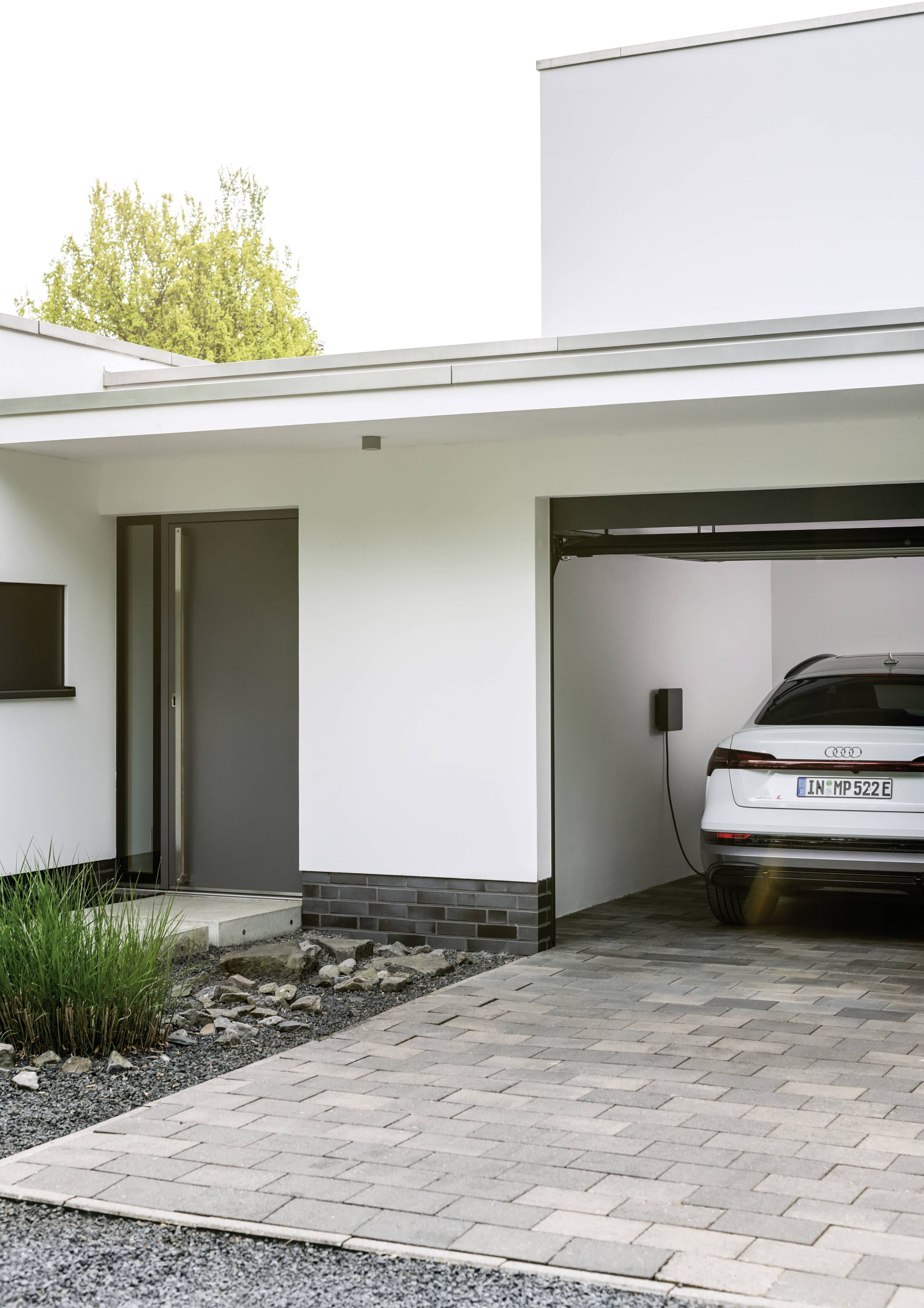 Ein modernes Elektroauto wird in einer offenen Garage aufgeladen. Das Haus hat weiße Wände und einen gepflasterten Hof.