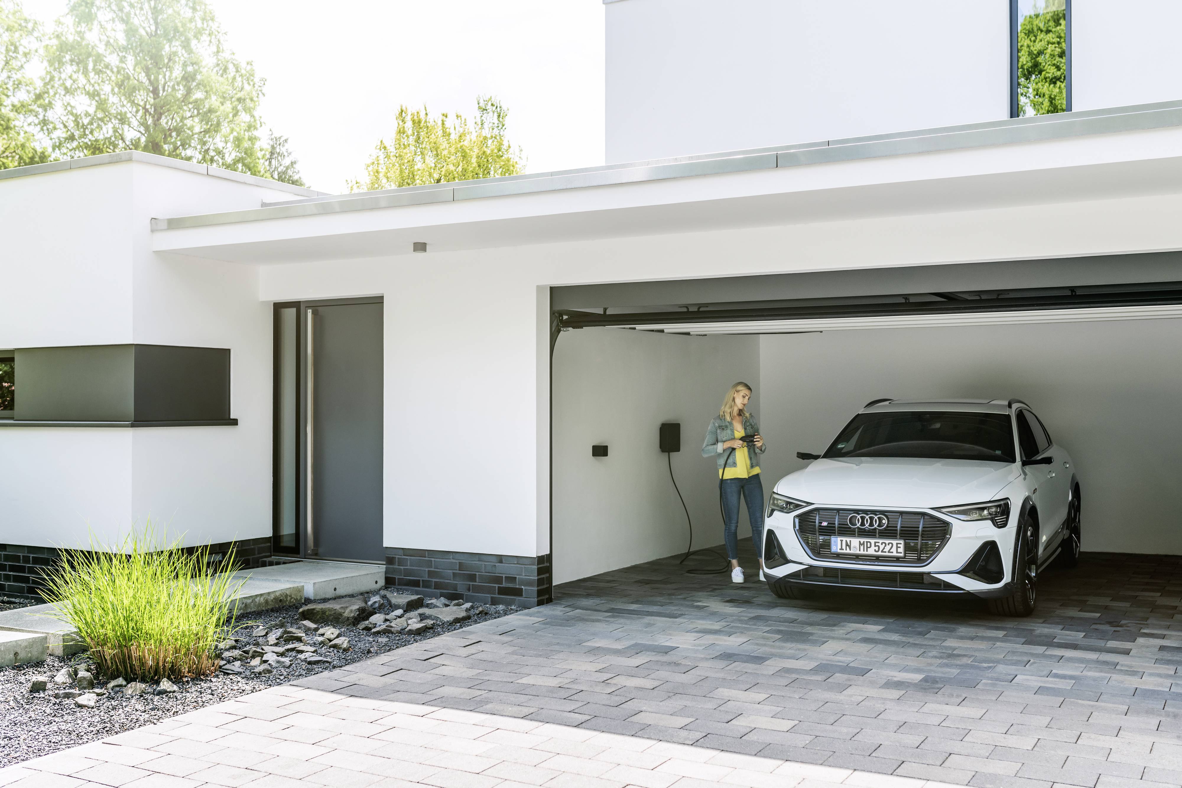 Eine Frau lädt ein Elektroauto in einer modernen Garage auf. Das Auto ist weiß und steht unter einem offenen Garagentor.