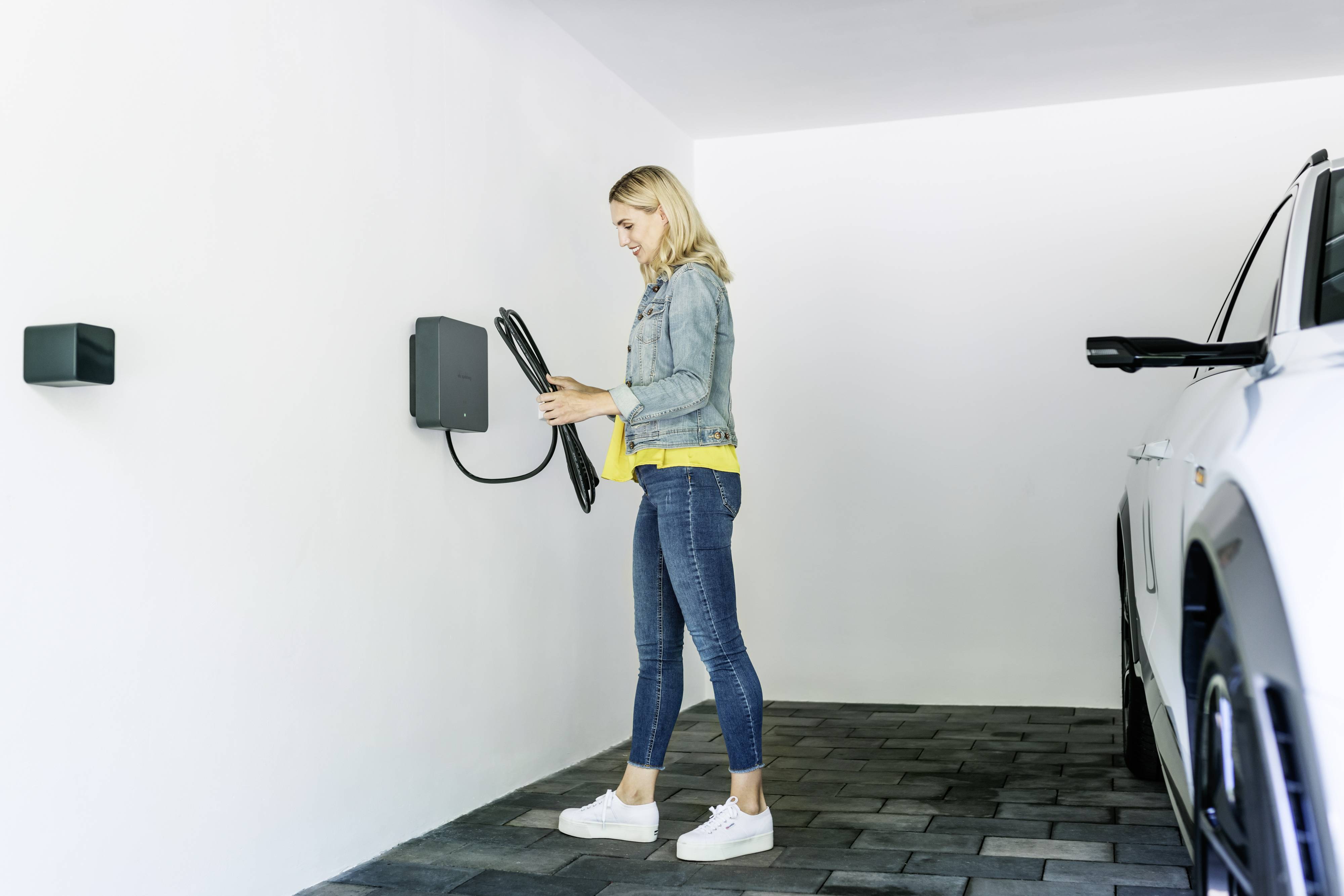 Eine Person steht in einer Garage und bedient eine Ladestation für ein Elektroauto. Ein Auto ist rechts im Bild zu sehen.