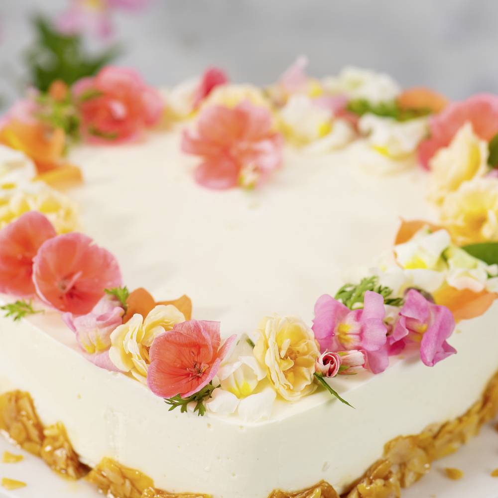 Ein quadratischer Kuchen mit weißem Zuckerguss, dekoriert mit bunten Blumen, auf einem Tisch mit verschwommenem Hintergrund.
