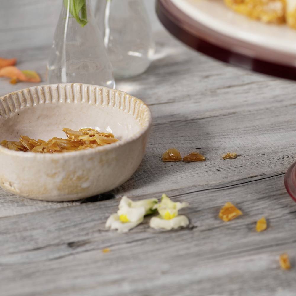 Kleine, flache Schale mit gebrochenen Karamellstücken auf Holztisch. Hintergrund zeigt eine Glasvase mit Blumen.