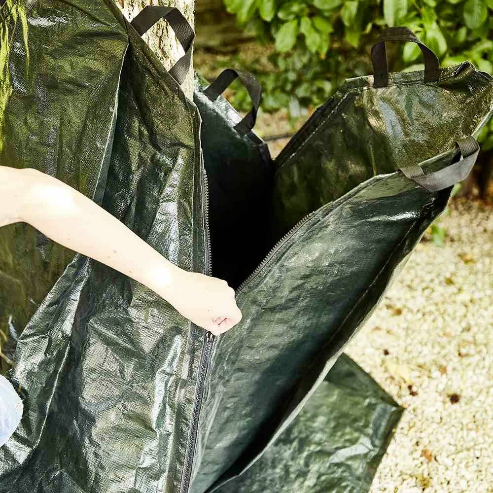 Eine Person öffnet einen großen, grünen Gartensack mit Reißverschluss. Im Hintergrund sind Bodengitter und Pflanzen zu sehen.