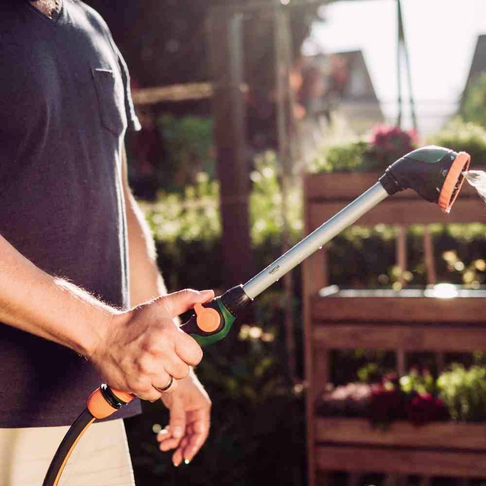 Eine Person gießt Pflanzen in einem Garten mit einem Gartenschlauch. Im Hintergrund sind Holzkästen mit Blumen zu sehen.