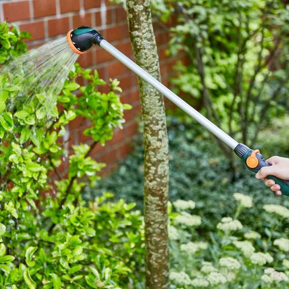 Eine Person bewässert einen Garten mit einem langen Schlauch. Pflanzen und ein Baum sind im Hintergrund zu sehen.