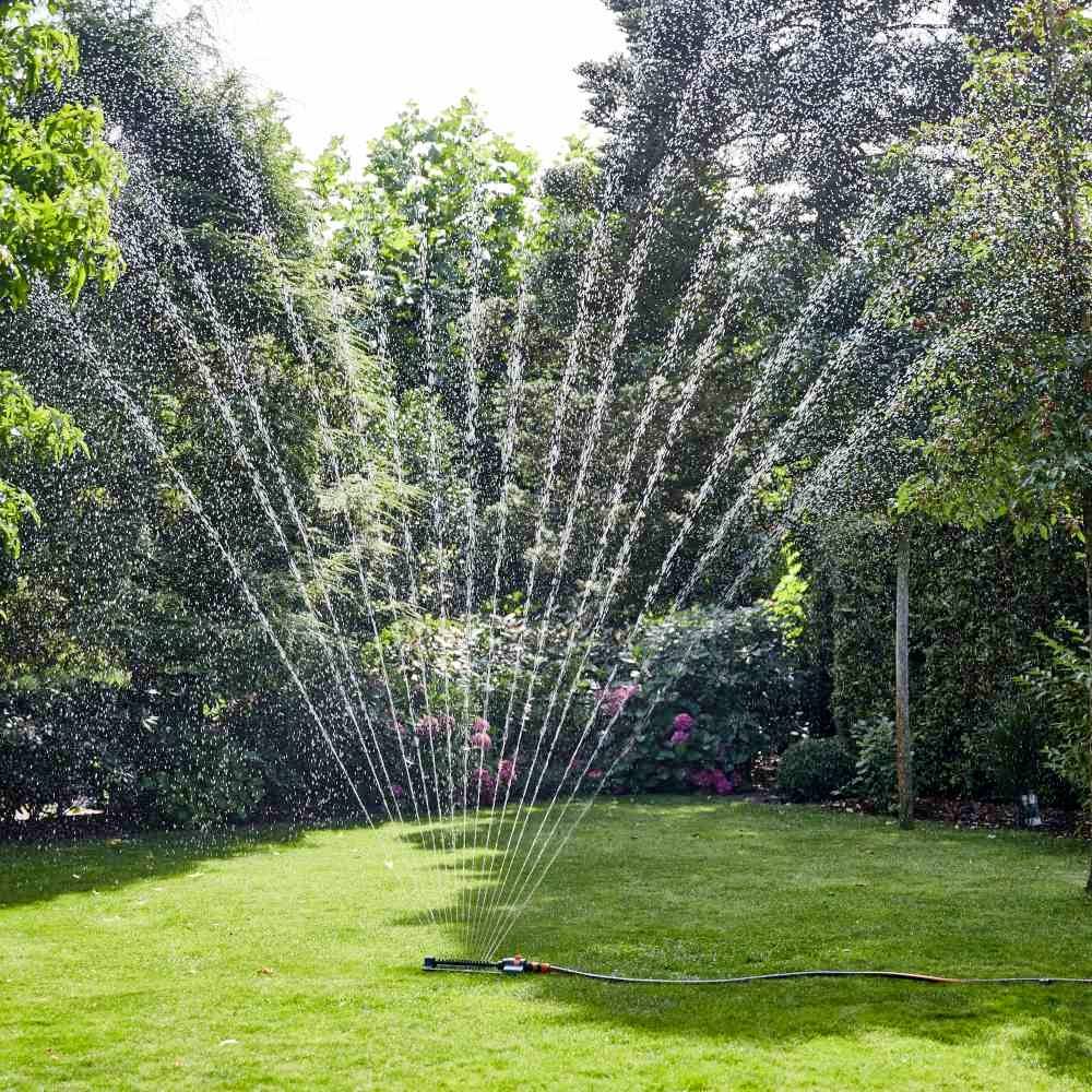 Eine grüne Rasenfläche wird von einem Rasensprenger bewässert, der Wasser in einem großen Fächer über das Gras sprüht. Im Hintergrund sind Büsche und Bäume.
