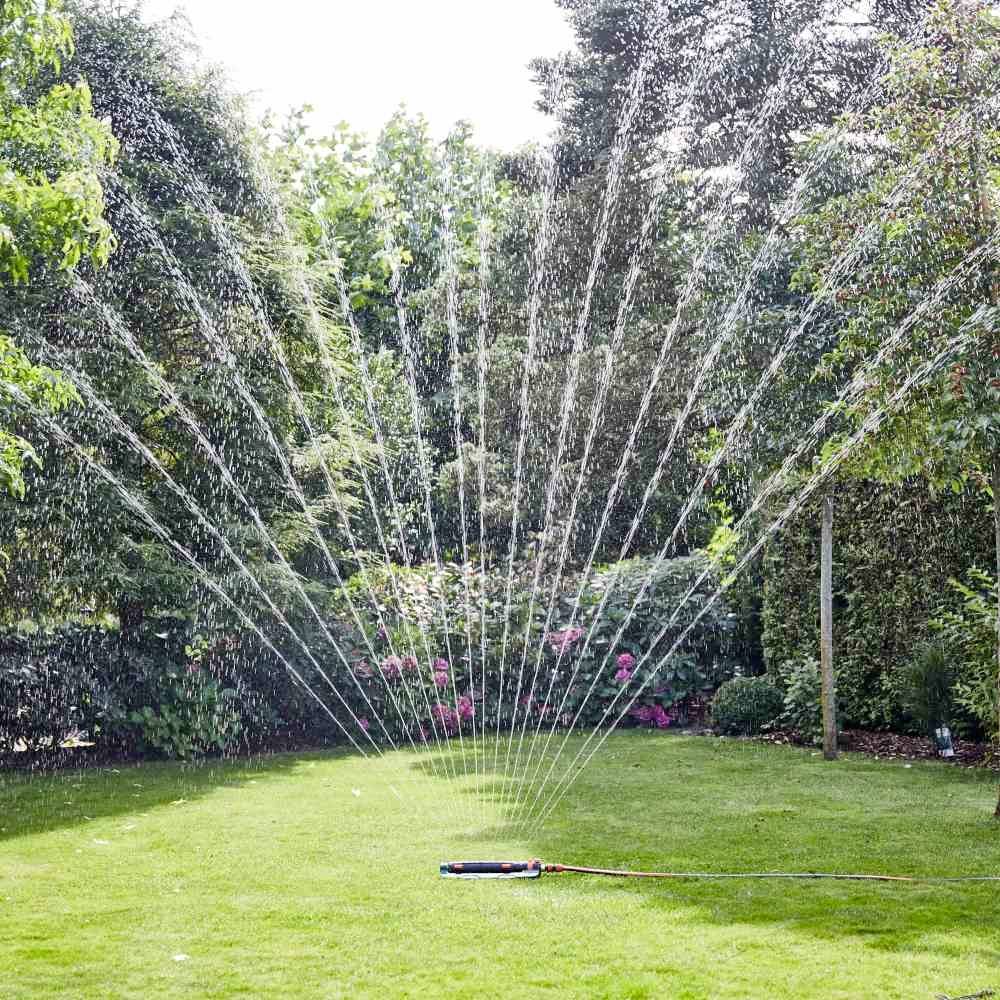 Ein Gartensprinkler bewässert Rasen mit Wasserstrahlen in einem Garten, umgeben von Bäumen und Blumen.