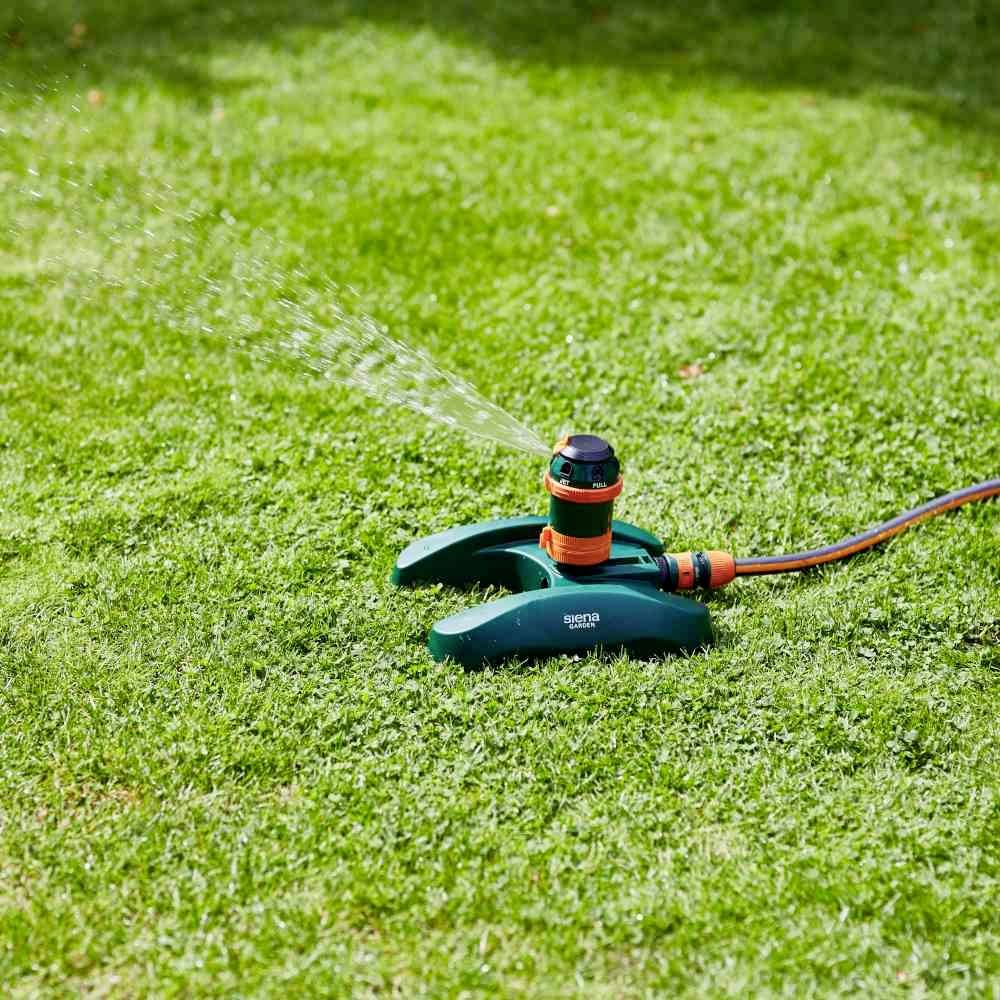 Ein Rasenregner besprüht gleichmäßig das grüne Gras einer Gartenfläche. Ein Wasserschlauch ist an den Regner angeschlossen.