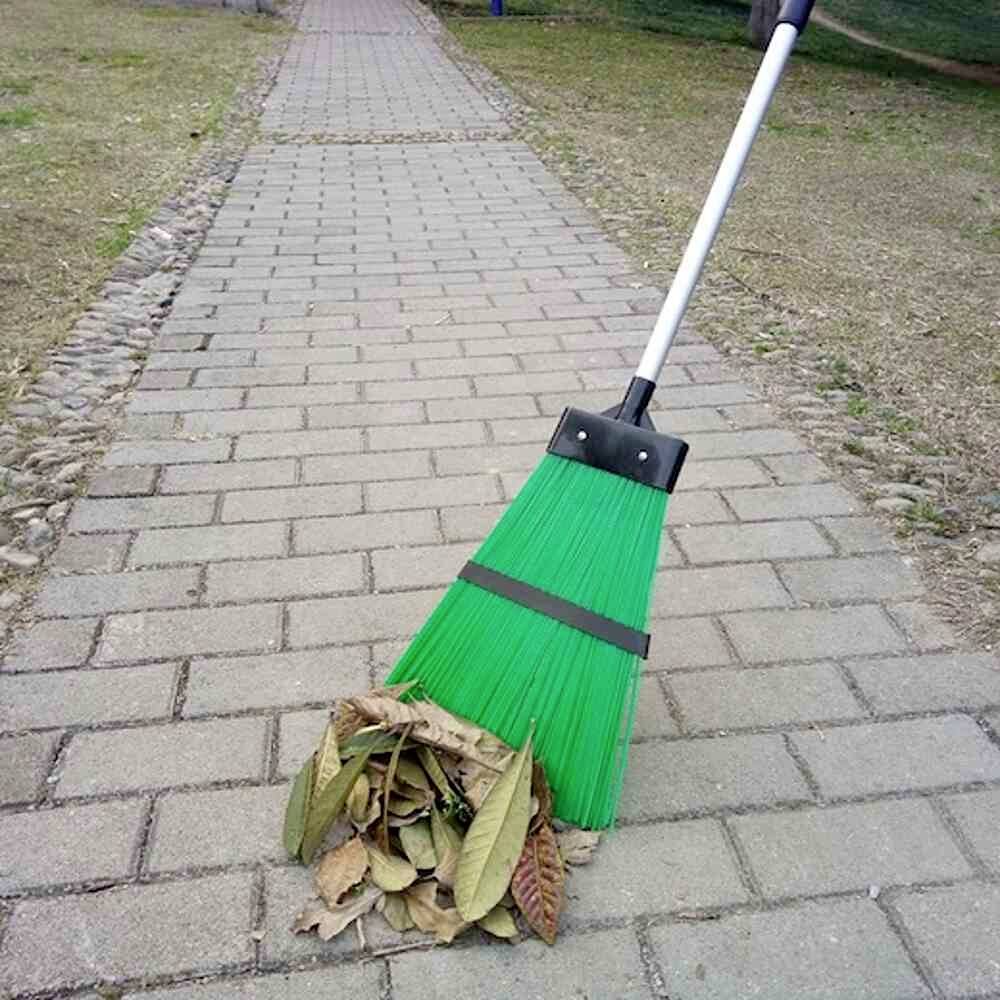 Ein grüner Besen fegt Blätter auf einem gepflasterten Gehweg. Seite des Weges ist grasbewachsen. Klarer, sonniger Tag.
