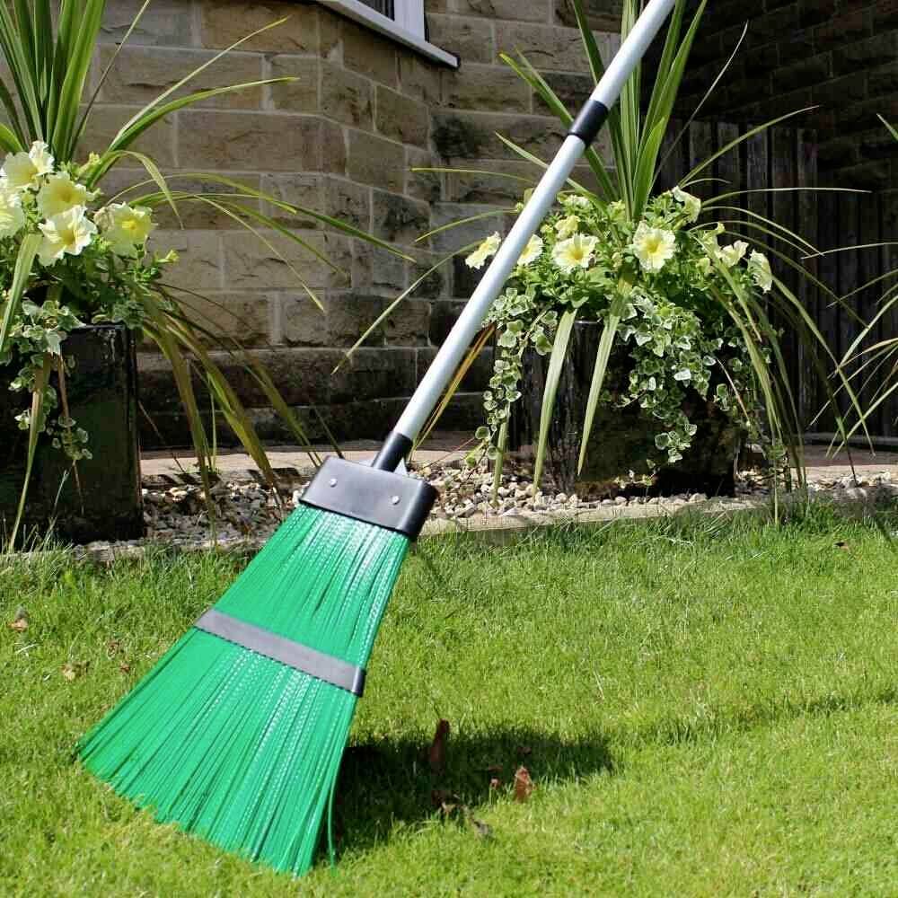 Ein grüner Besen lehnt draußen gegen eine Hauswand, umgeben von zwei Blumentöpfen mit gelben Blumen und grünen Blättern.