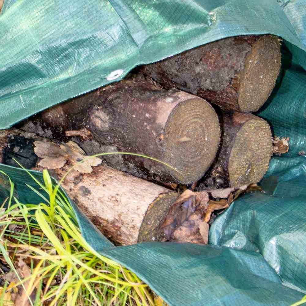 Holzscheite liegen unter einer grünen Plane auf dem Boden. Ein paar Blätter liegen daneben, umgeben von Gras.
