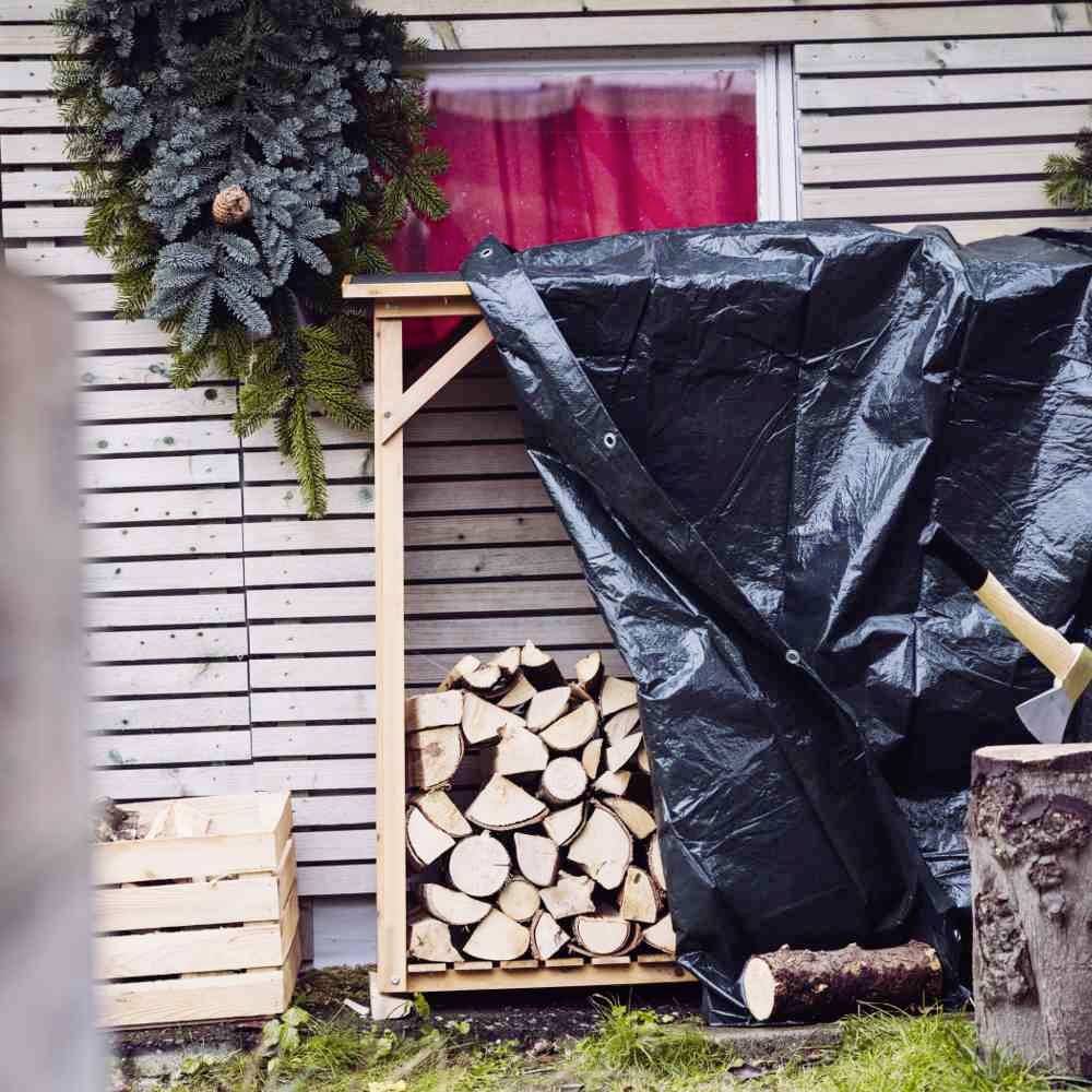 Holzstapel unter schwarzer Plane neben stapelbare Holzbox, daneben grauer Kranz an Holzwand mit roten Vorhängen im Hintergrund.