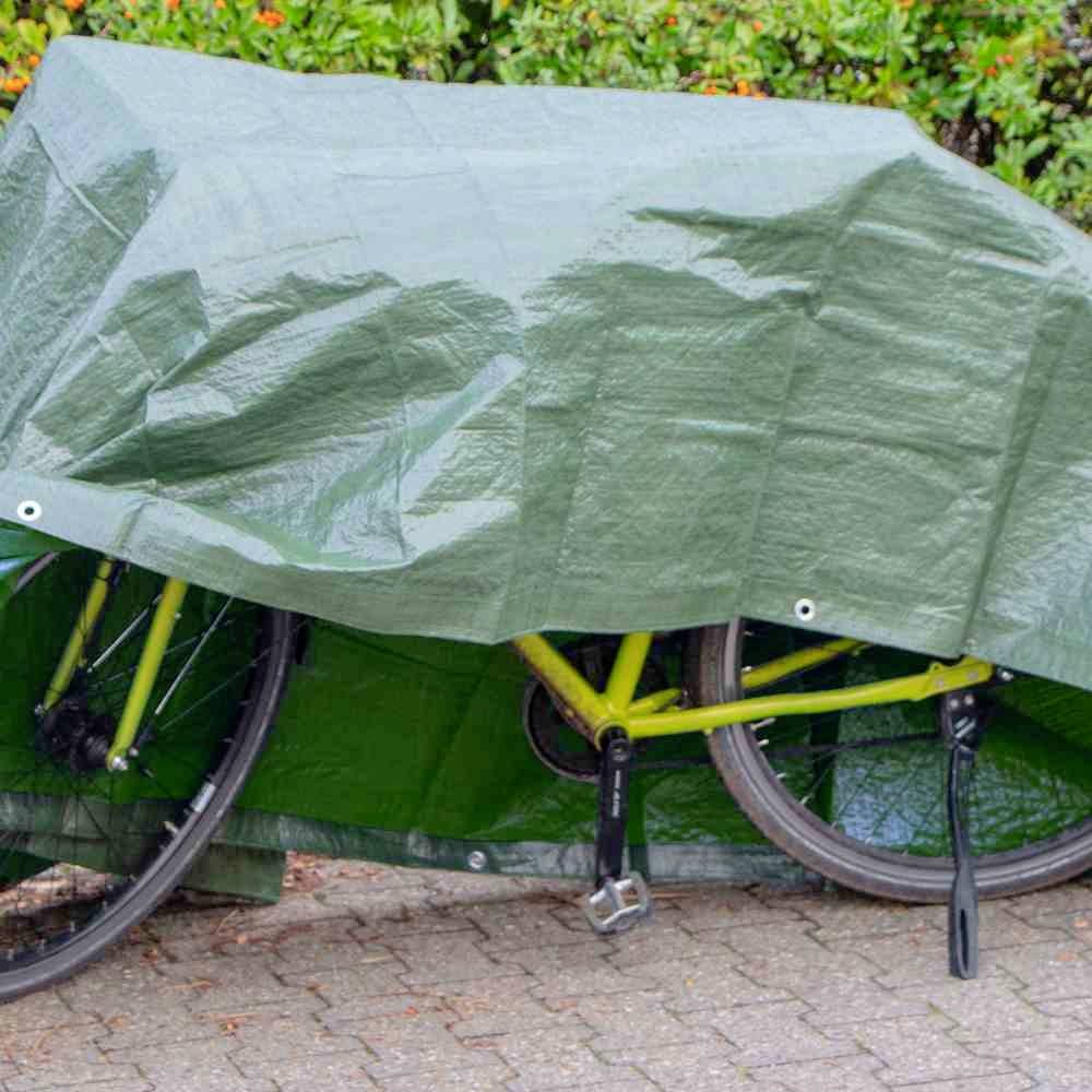 Ein grünes Fahrrad steht draußen auf einem Gehweg, teilweise mit einer grünen Plane abgedeckt. Im Hintergrund sind Büsche zu sehen.