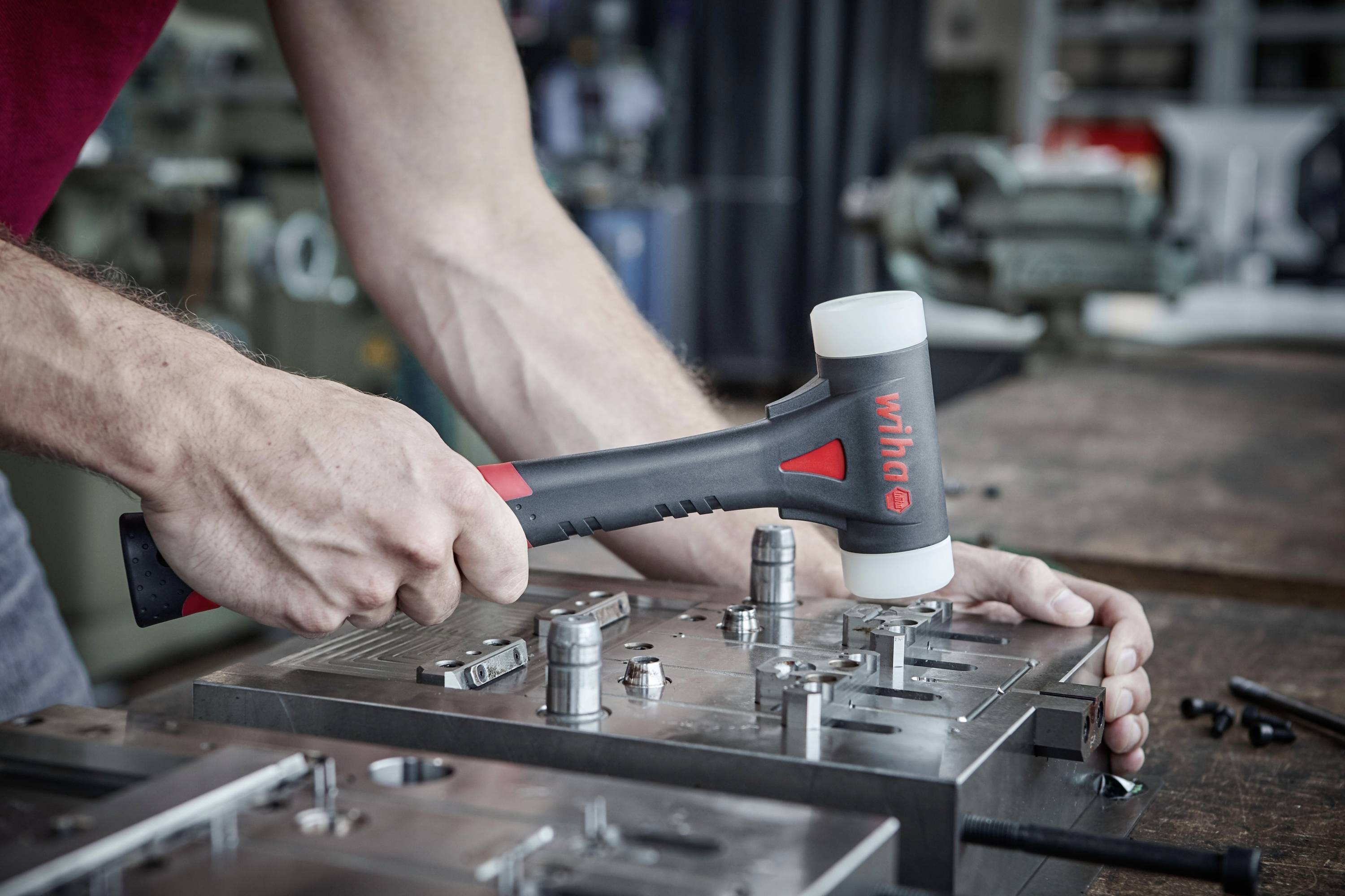 Eine Person bearbeitet mit einem Gummihammer eine Metallform. Der Hammer hat einen schwarzen Griff mit rotem 'wiha'-Logo. Im Hintergrund stehen Maschinen.