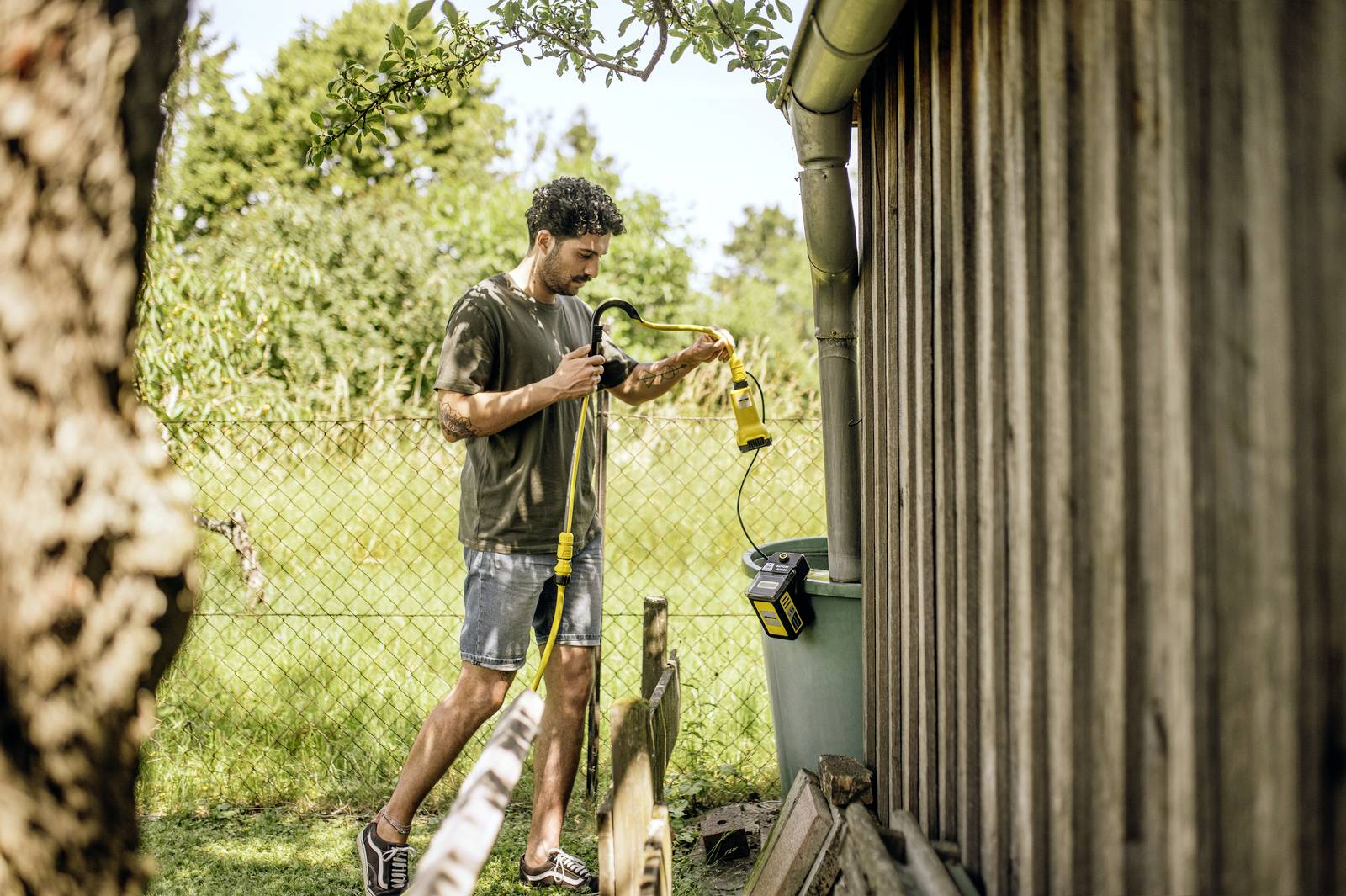 Mann bewässert Gartenpflanzen mit gelbem Gartenschlauch aus Wassertonne neben Holzwand, grüne Landschaft im Hintergrund.
