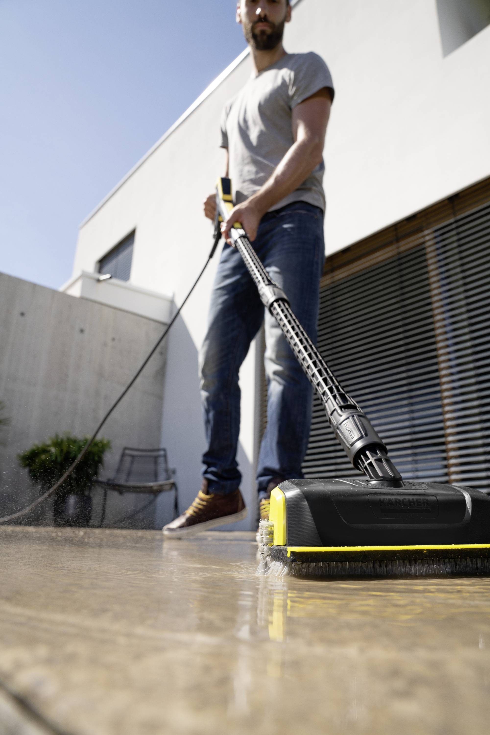 Ein Mann reinigt eine Terrasse mit einem Hochdruckreiniger. Im Hintergrund ist ein modernes Gebäude mit großen Fenstern sichtbar.