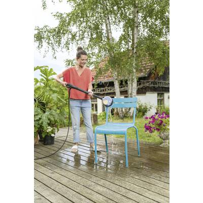Eine Frau spritzt auf einer Terrasse einen blauen Stuhl mit einem Wasserschlauch ab. Im Hintergrund steht ein Haus mit Bäumen.