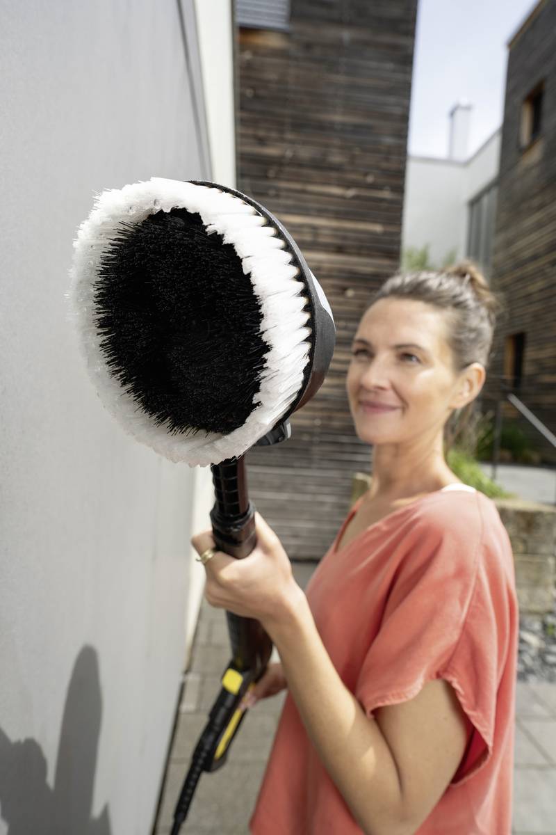Eine Frau in einem orangefarbenen Oberteil hält eine große Reinigungsbürste in der Hand und lächelt, während sie draußen steht.