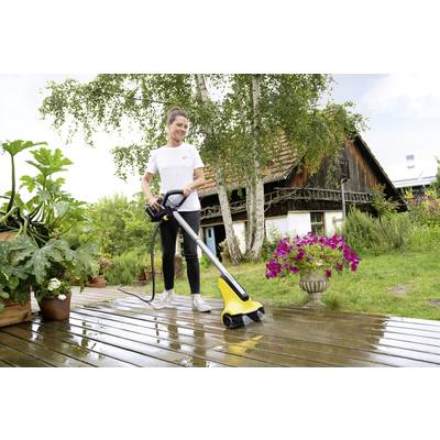 Eine Person reinigt eine Holzterrasse mit einem elektrischen Reinigungsgerät. Im Hintergrund ist ein Haus mit Garten und Blumentöpfen zu sehen.