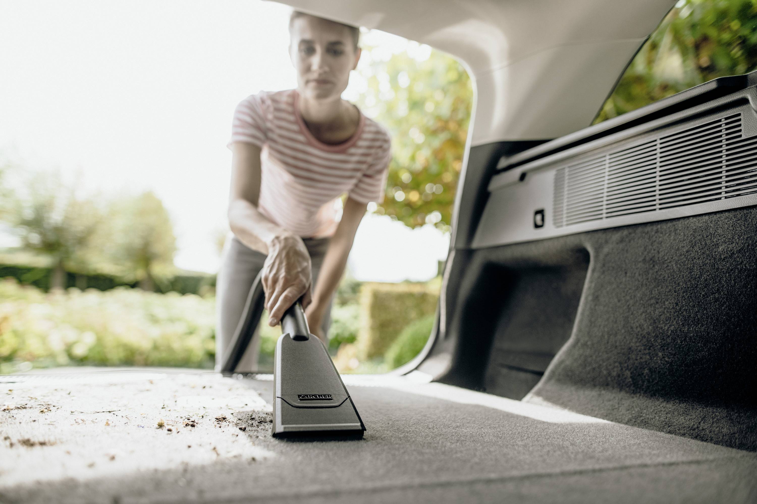 Eine Person saugt den Kofferraum eines Autos aus, um ihn zu reinigen. Sie verwendet einen kleinen Handstaubsauger im Sonnenschein.