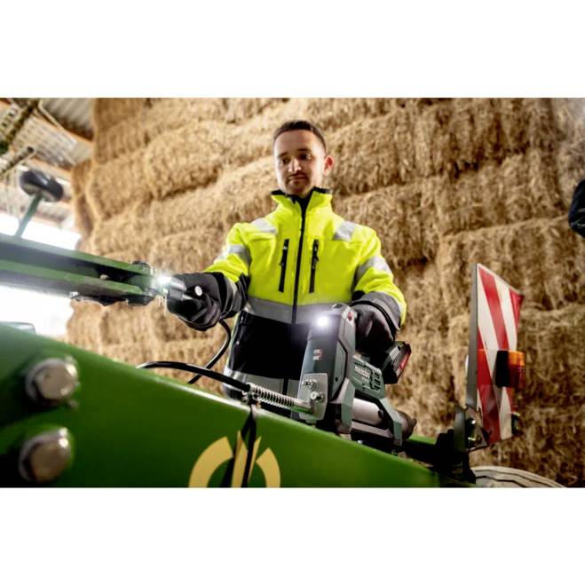 Eine Person in einer Warnweste bedient Maschinen in einer Scheune, die mit gestapelten Heuballen gefüllt ist.