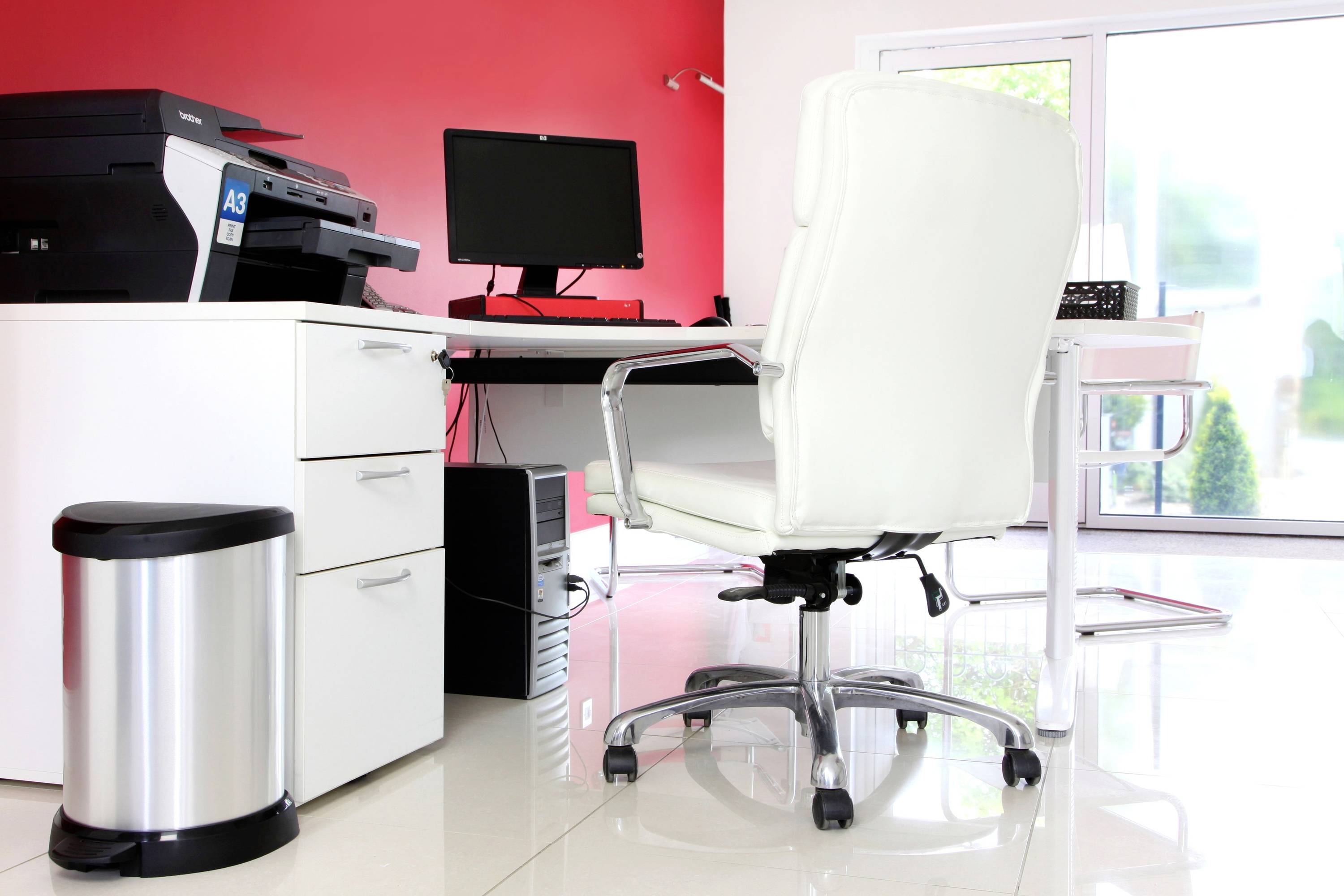 Bürozimmer mit weißem Schreibtisch, weißem Bürostuhl, schwarzem Computerbildschirm und Drucker, roter Wand im Hintergrund.
