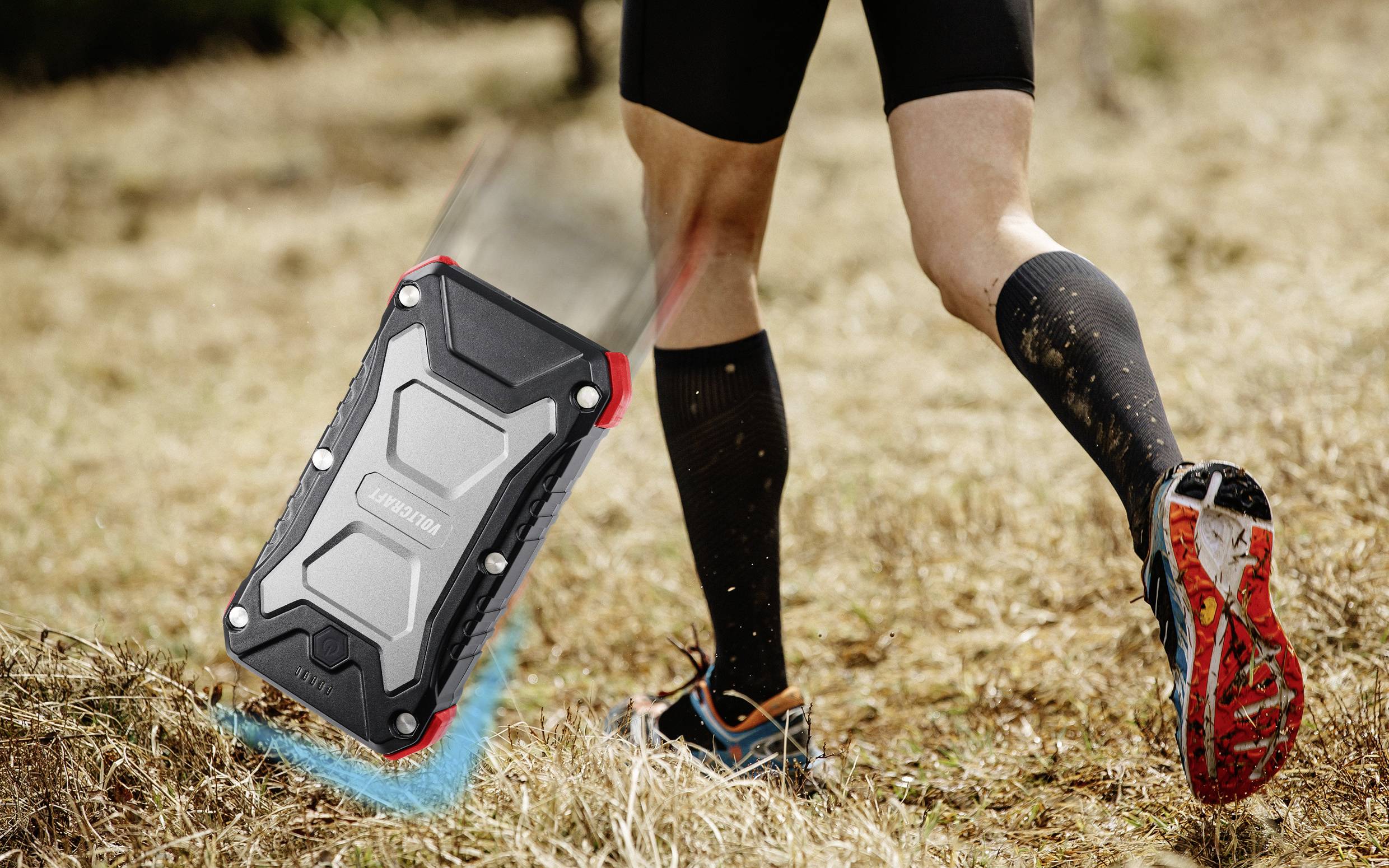 Ein Läufer in Sportuniform joggt im Freien auf einem unebenen Pfad. Im Vordergrund fällt ein robustes, stoßfestes Gerät auf den Boden.