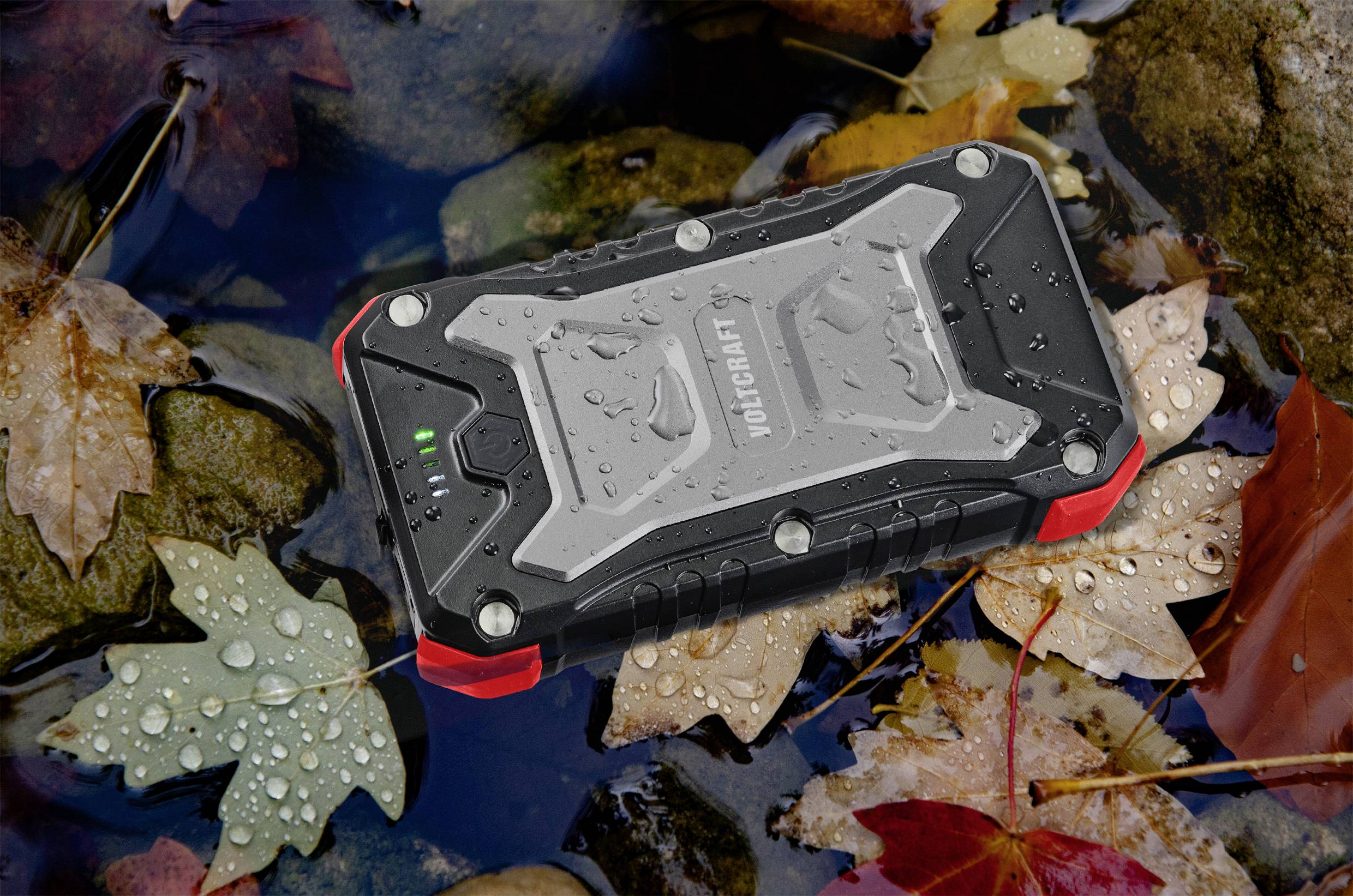 Eine wasserdichte Outdoor-Powerbank liegt in einer Pfütze, umgeben von herbstlichen Blättern. Regentropfen sind auf der Oberfläche sichtbar.